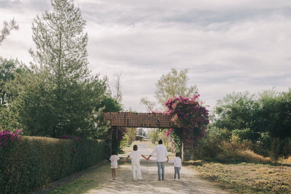 People walking down a garden path beneath a rustic wooden archway covered in pink bougainvillea on a cloudy day.