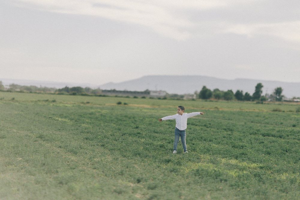 A person stands with outstretched arms in a vast green meadow with mountains in the distant background.