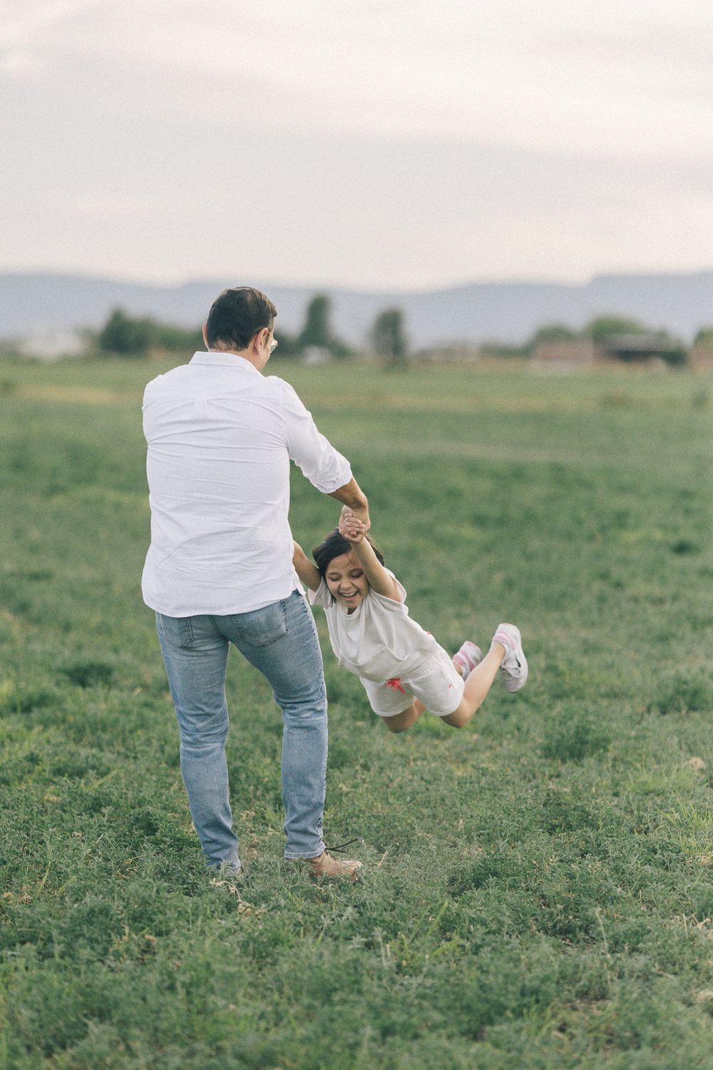 Adult in white shirt playfully swinging child in open grassy field with mountains in background.