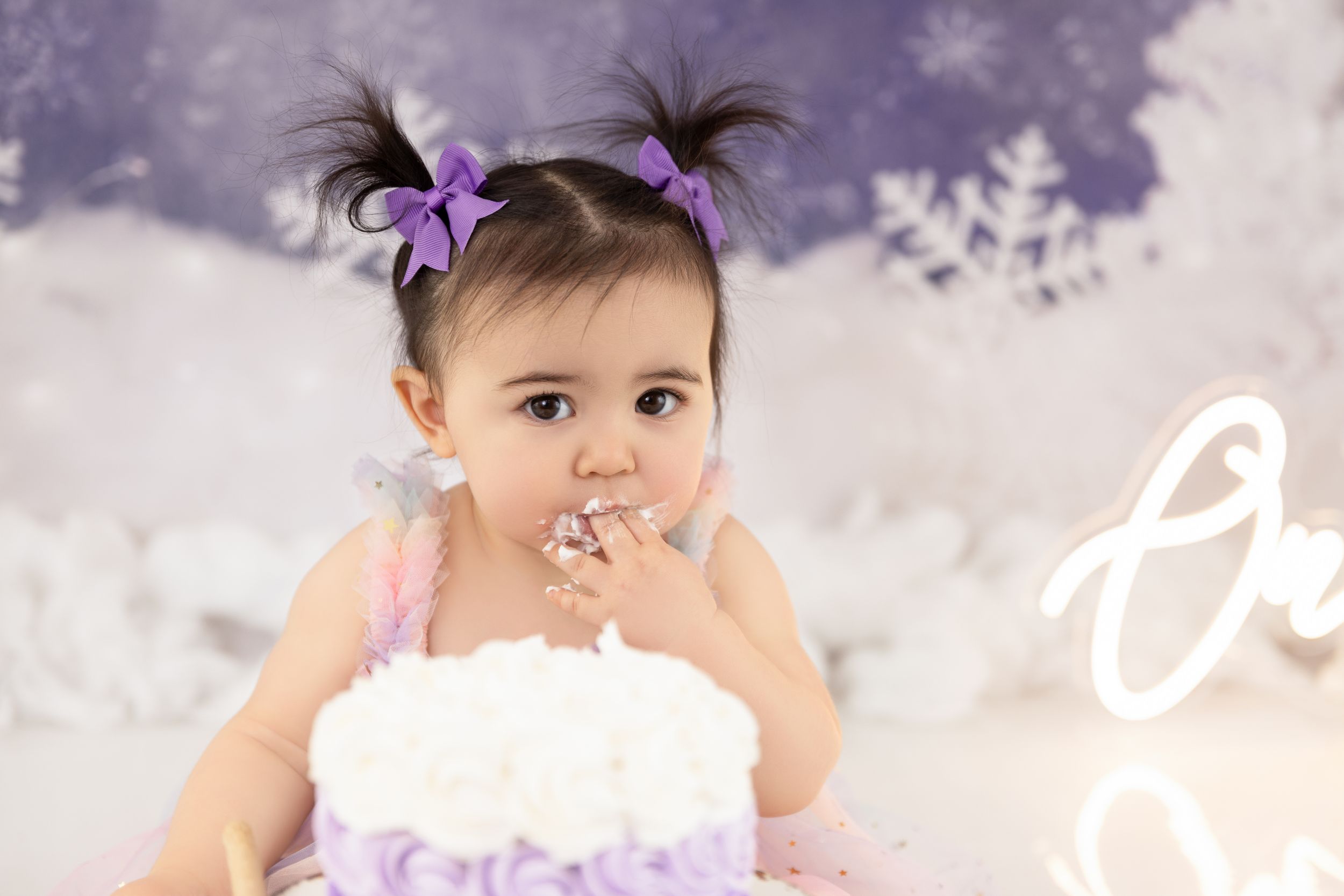 Small child in pink outfit enjoying cake against purple winter backdrop.