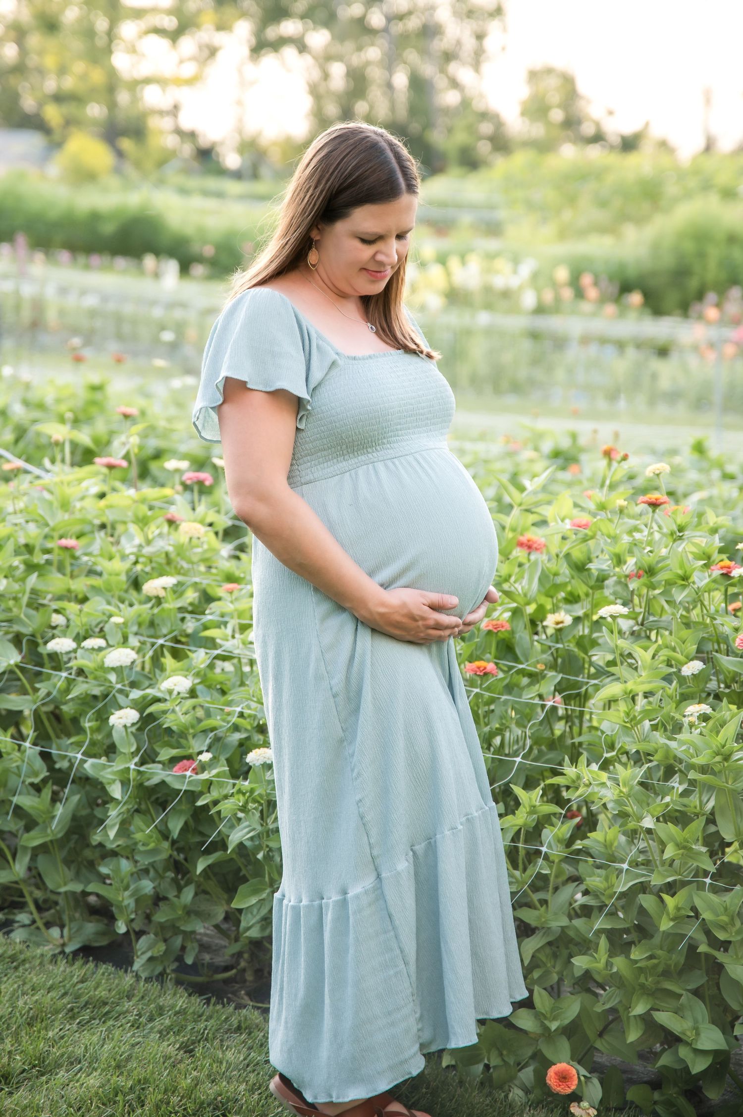 Pregnant woman in light blue maxi dress stands in garden with blooming flowers during golden hour sunset.
