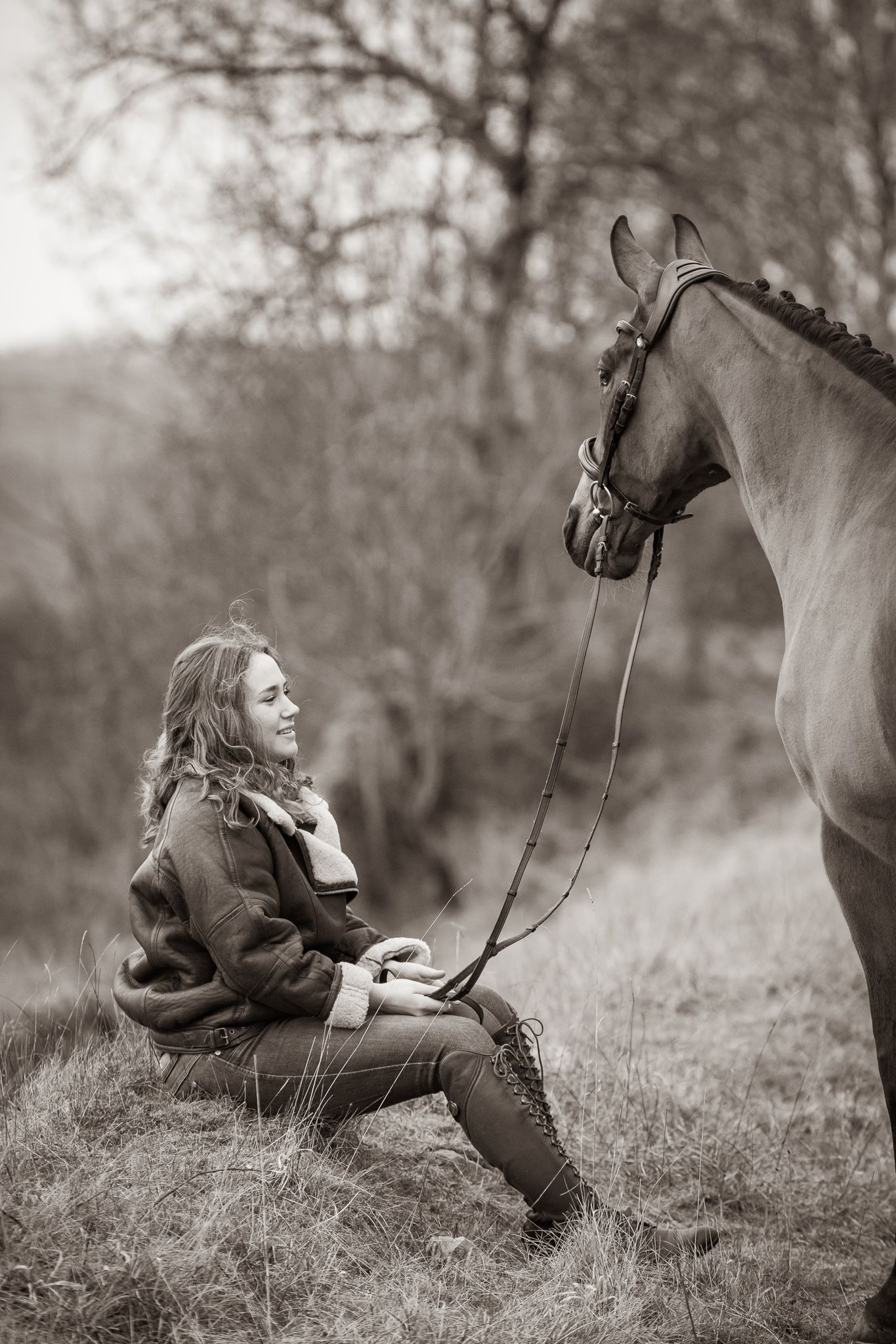A serene black and white portrait of someone in a flowy dress standing with a majestic white horse in a pastoral setting.