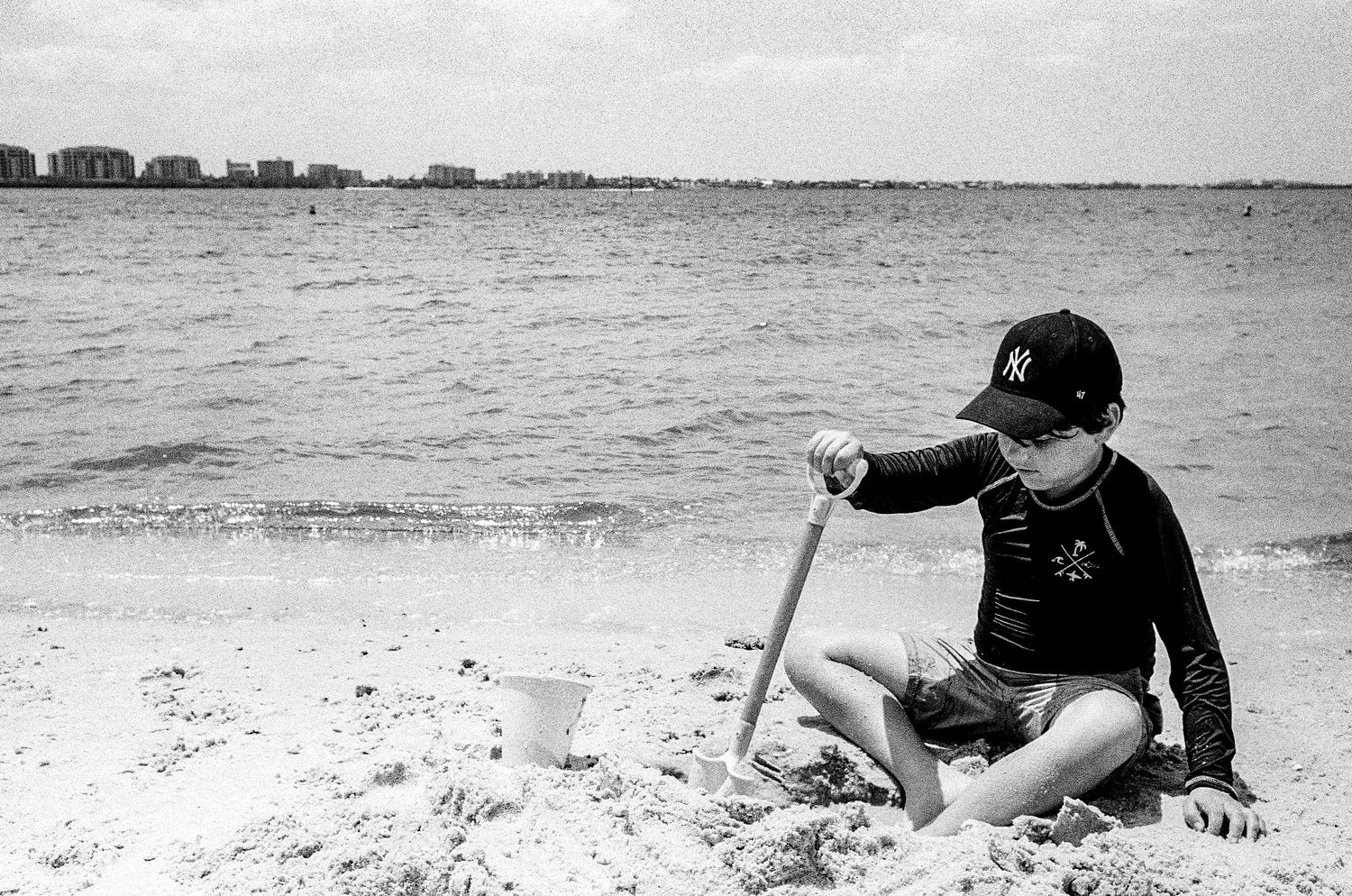 A child in black sits on a sandy beach using a shovel near a white bucket on a cloudy day.
