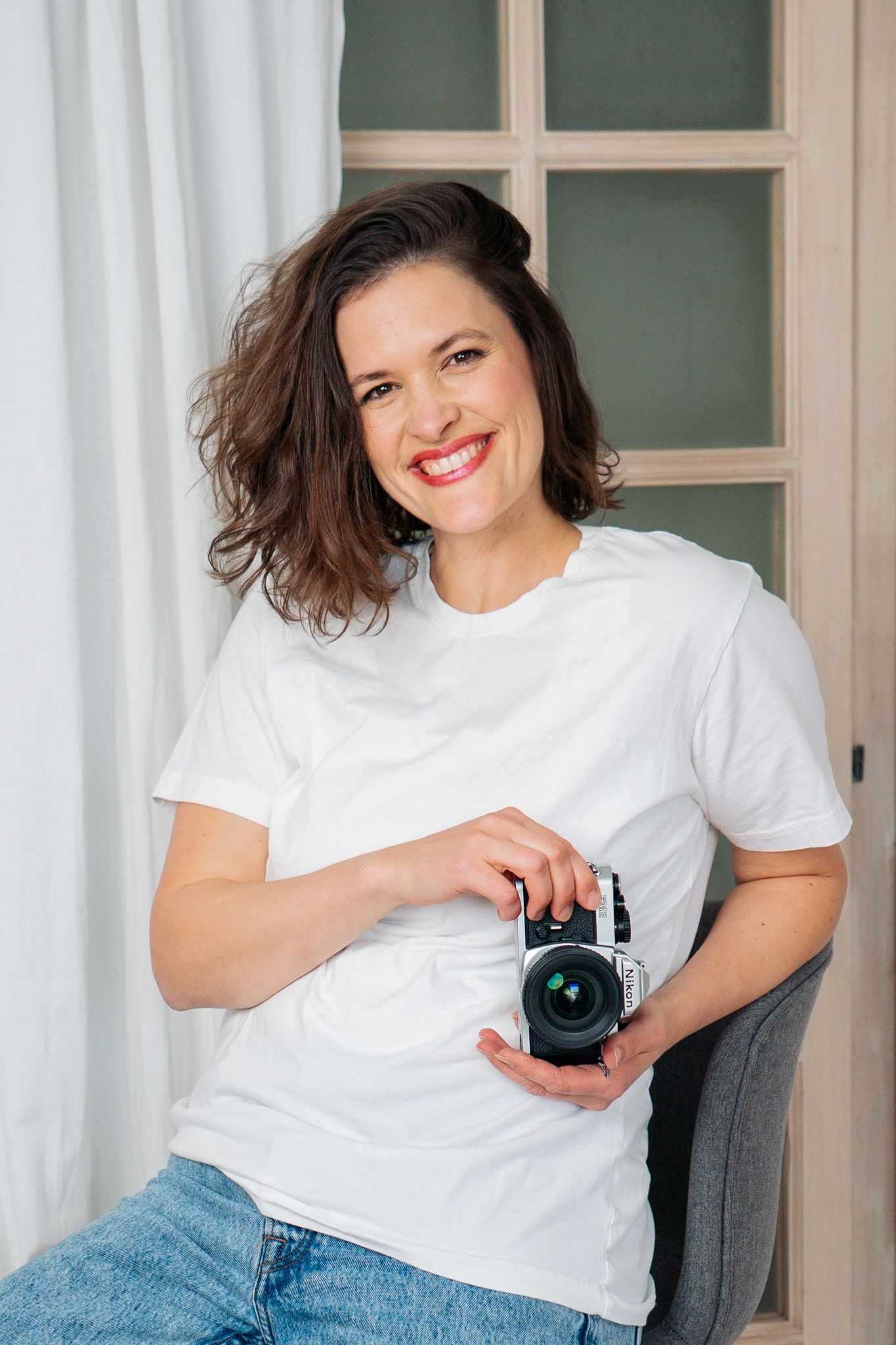 Casual portrait of photographer holding camera while sitting in bright interior setting.
