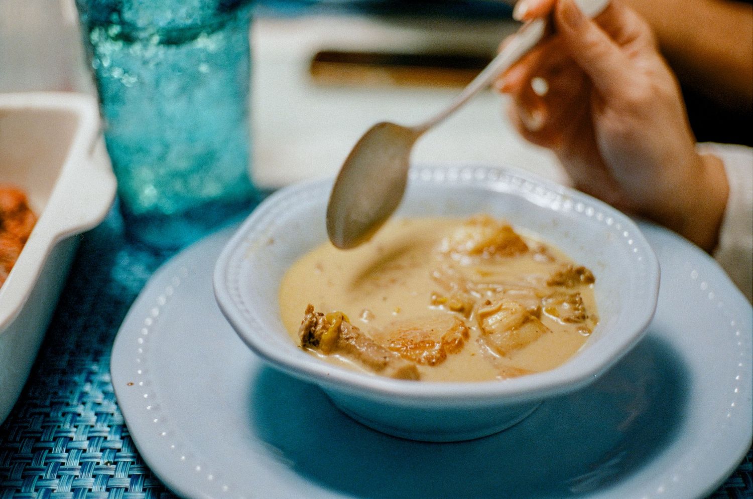 A spoonful hovers over a creamy white soup garnished with crispy toppings in a white bowl on a blue tablecloth.