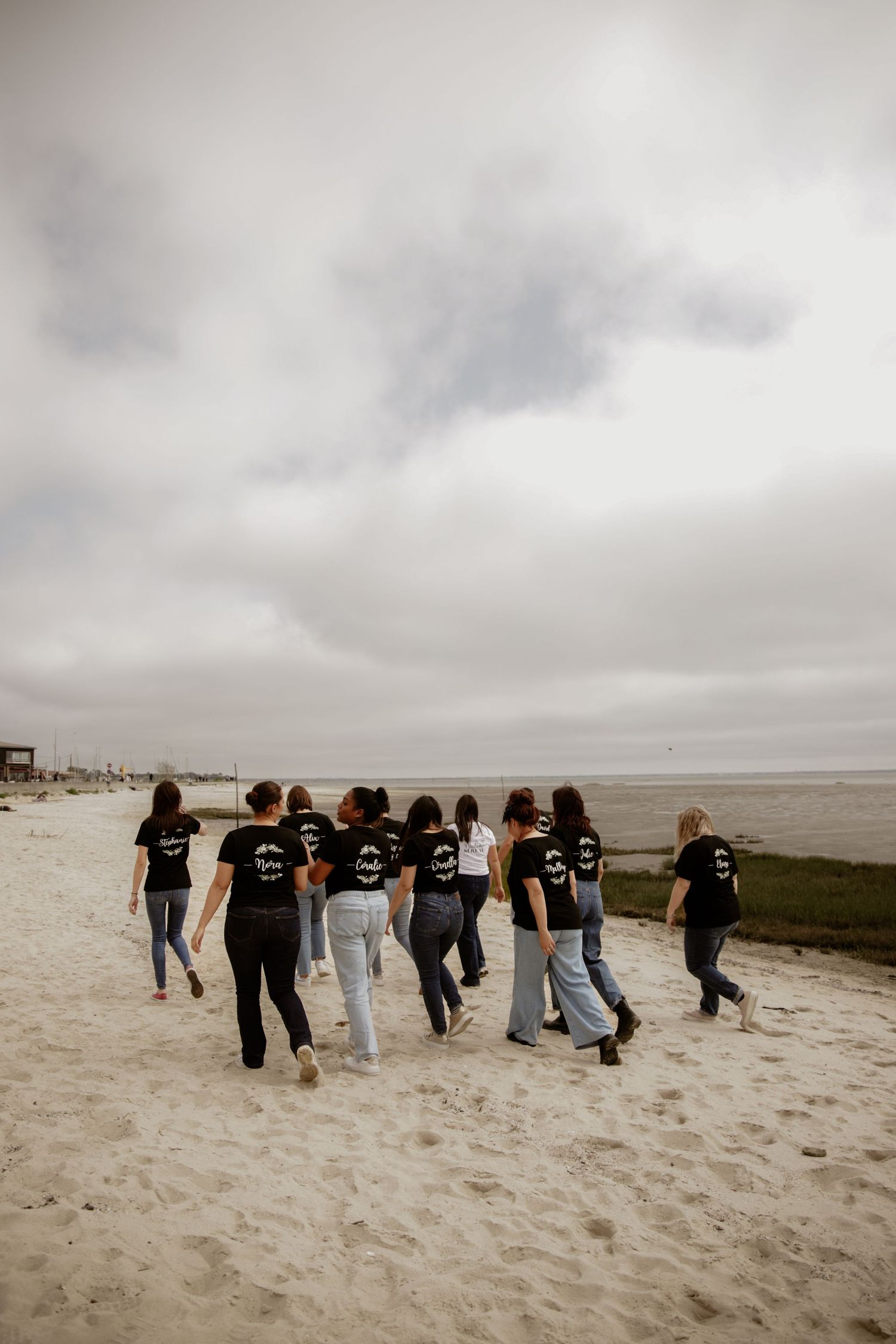 Group of friends wearing matching black shirts walk together along a cloudy beach shoreline.