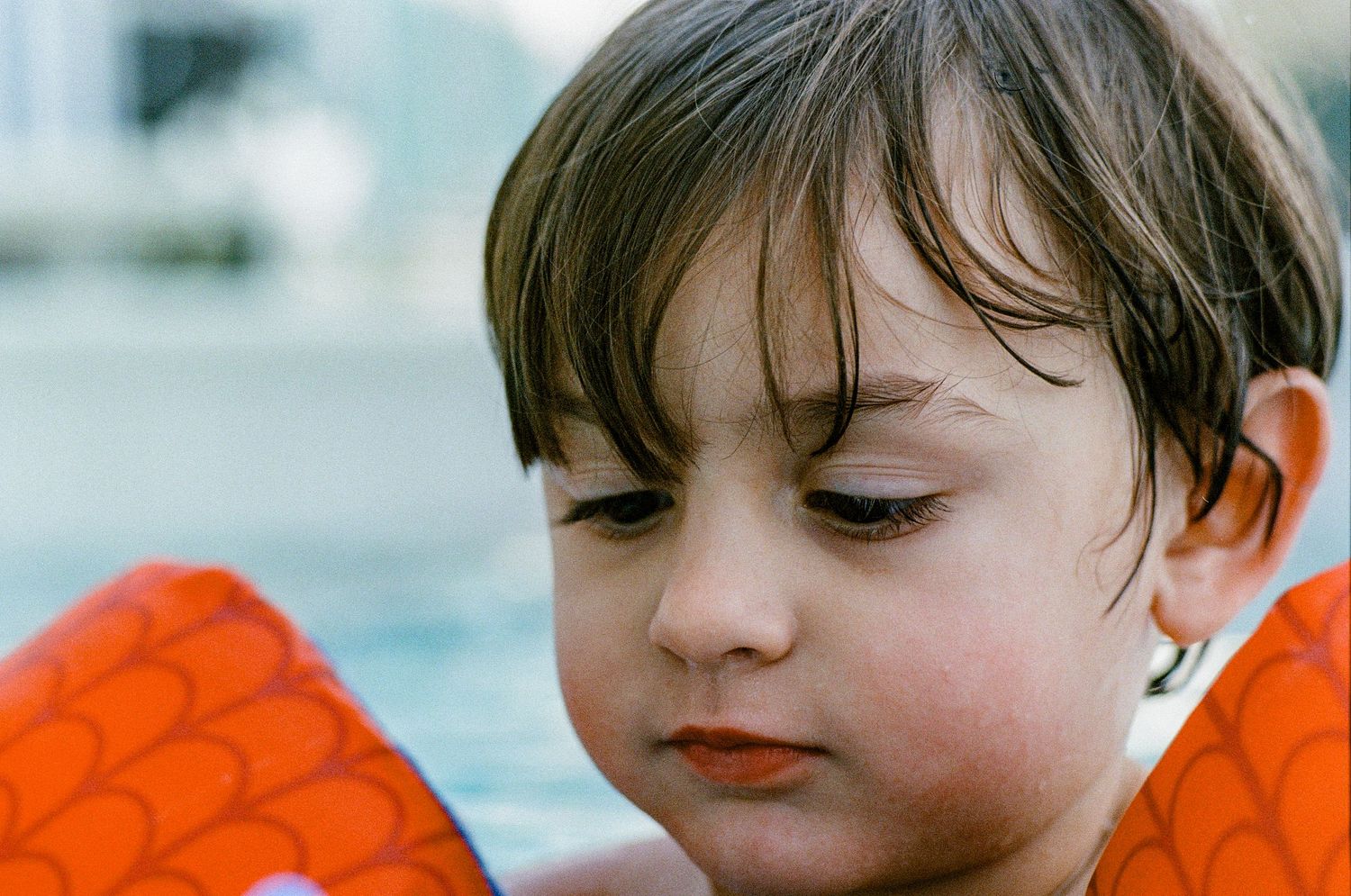 A child wearing orange water wings gazes downward with a pensive expression and dark bangs.