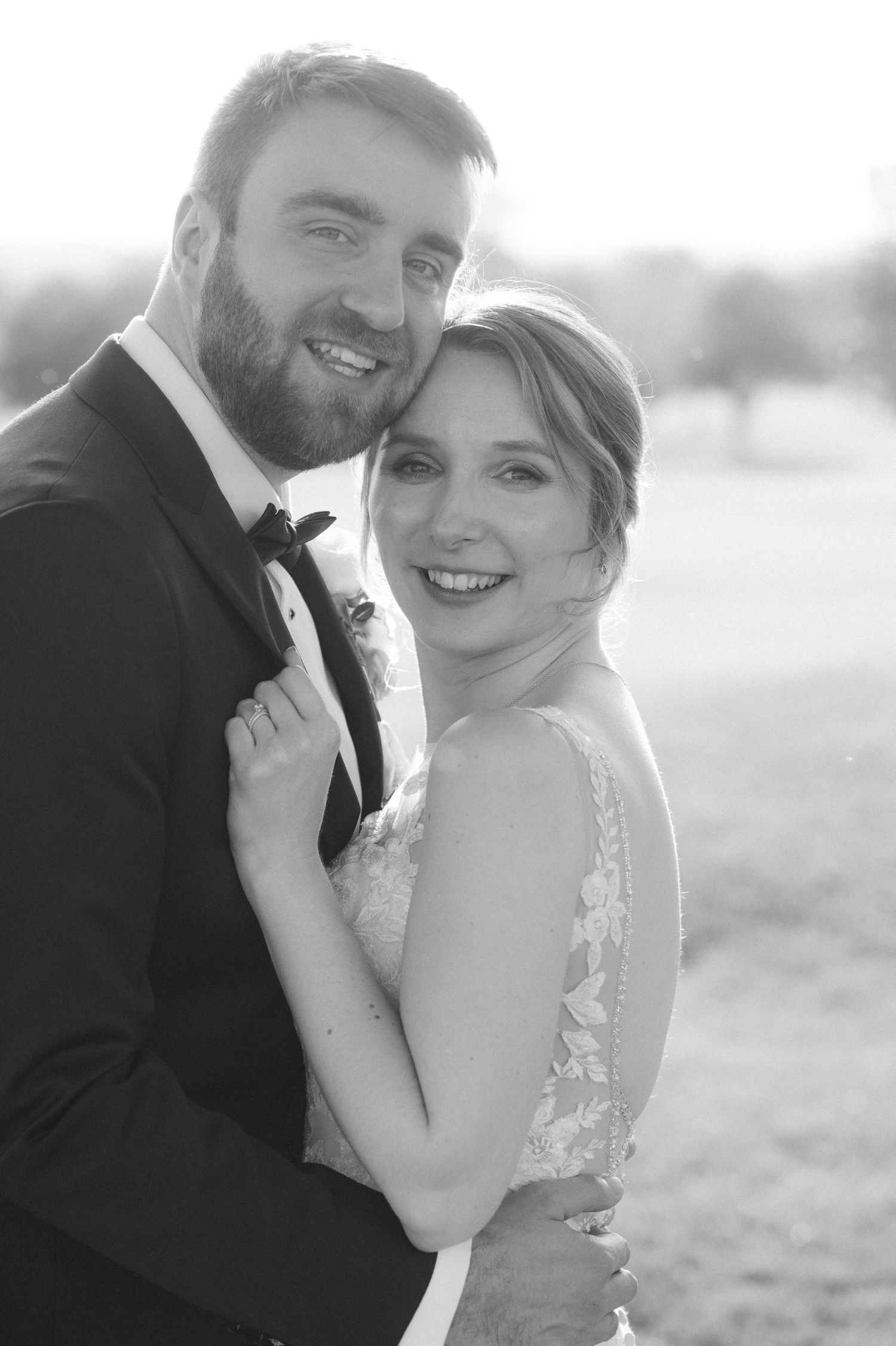 Black and white wedding portrait of smiling bride and bearded groom embracing outdoors in formal attire.