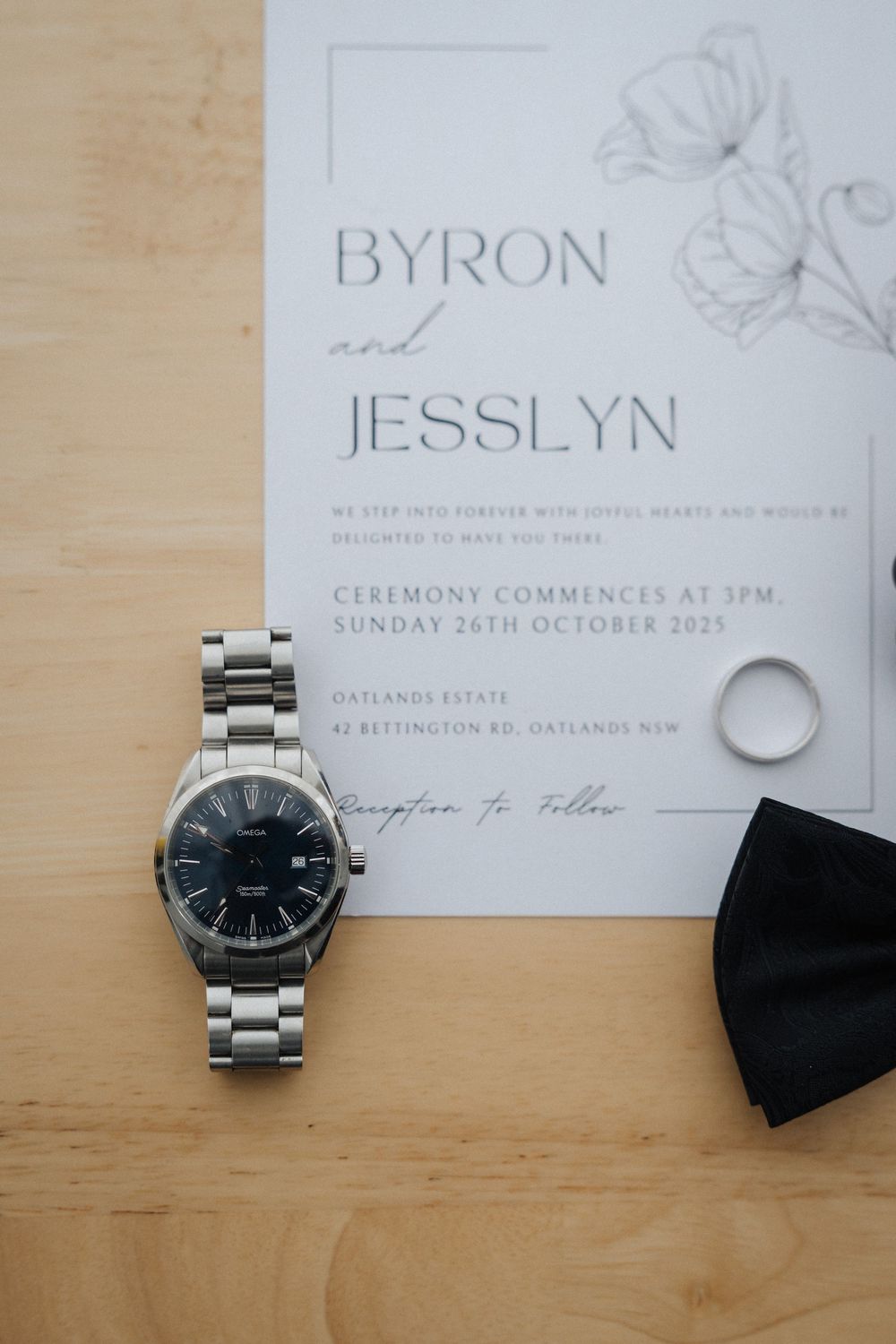 Wedding invitation for Byron and Jesslyn with a silver watch, gold ring, and black bow tie on a wooden surface.