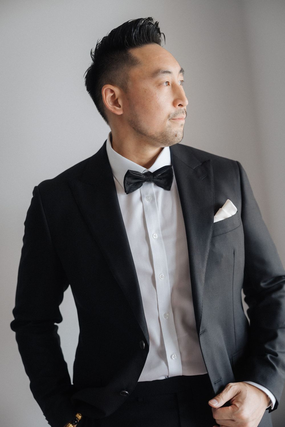 A well-dressed Asian man in a black tuxedo with bow tie and pocket square gazes confidently to the side.