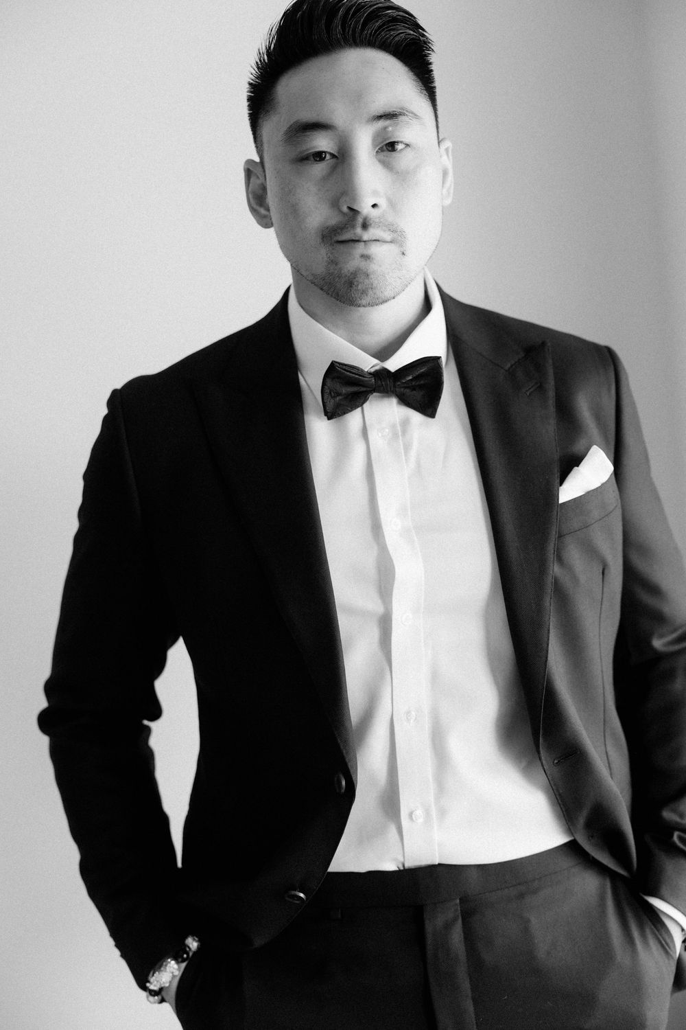Black and white portrait of a man in a tuxedo with bow tie and pocket square, hands in pockets.