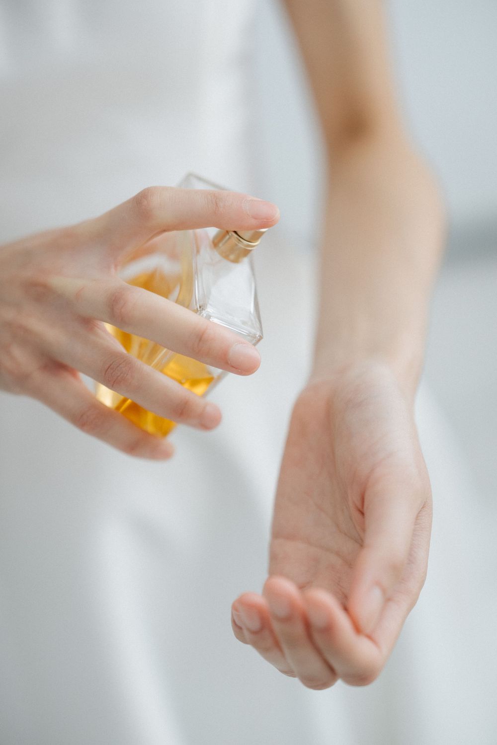 A woman spraying golden perfume from a glass bottle onto her wrist while wearing a white outfit.