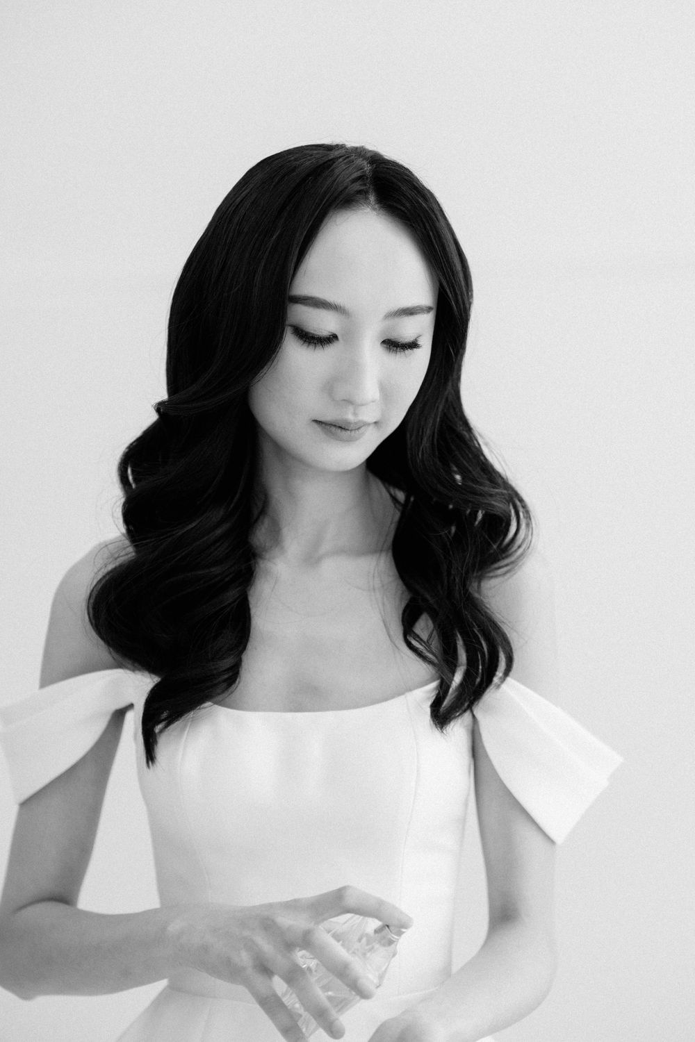 Black and white portrait of an elegant woman in white off-shoulder dress with wavy dark hair looking down.