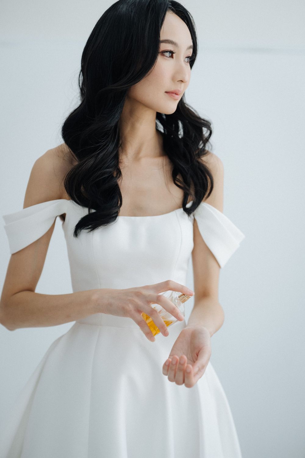 Elegant woman in white off-shoulder dress applying perfume, with long black wavy hair, against a light background.