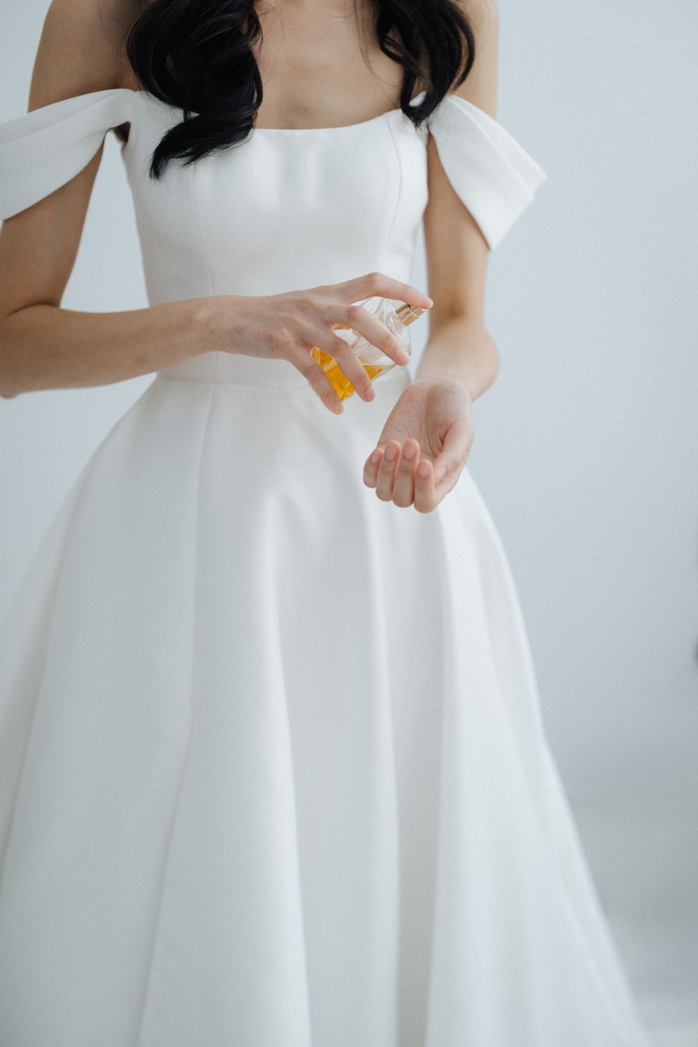 Bride in elegant off-shoulder white ball gown applying perfume to her wrist during wedding preparations.