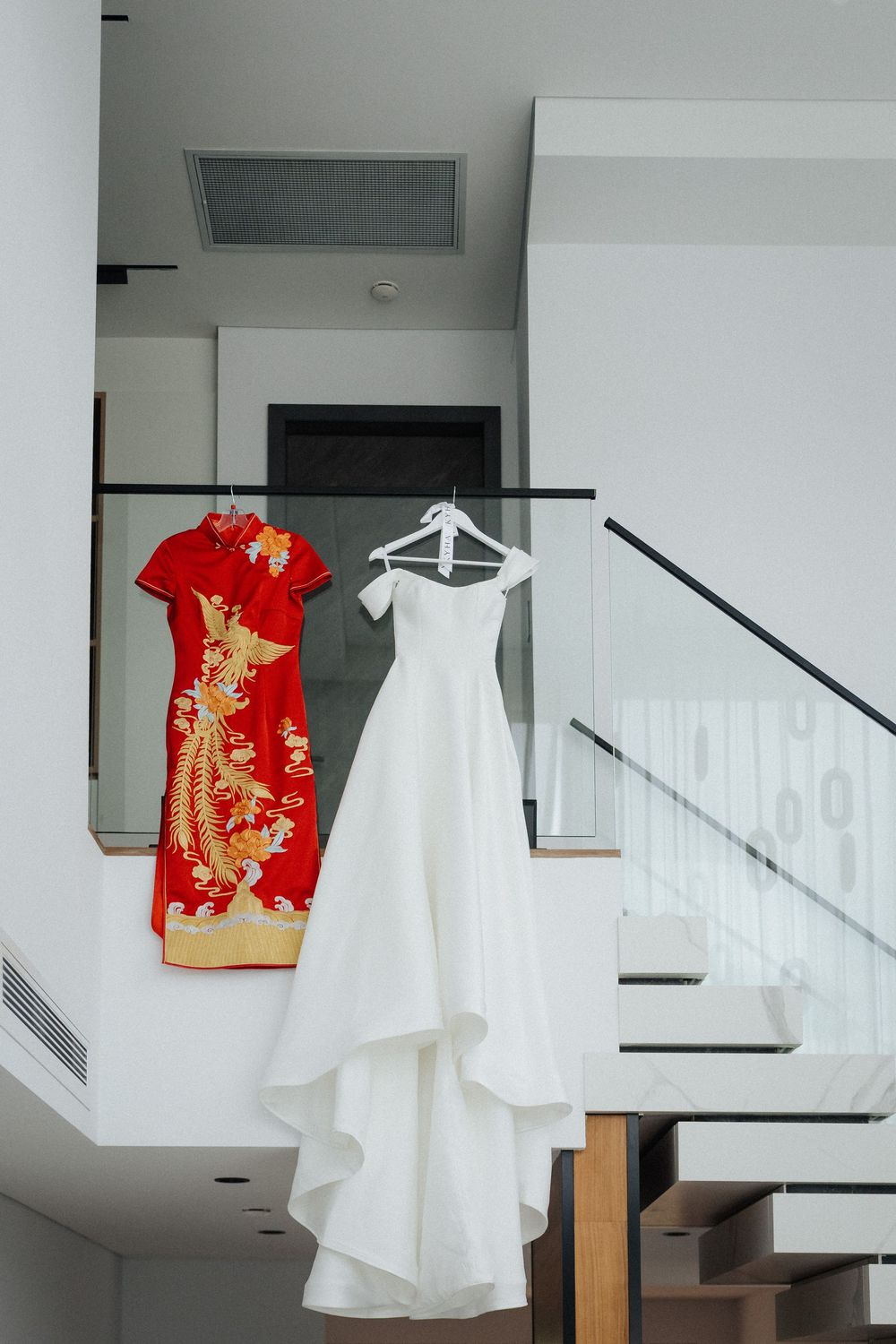 White wedding gown and red Chinese qipao dress hanging on staircase railing in modern white interior.