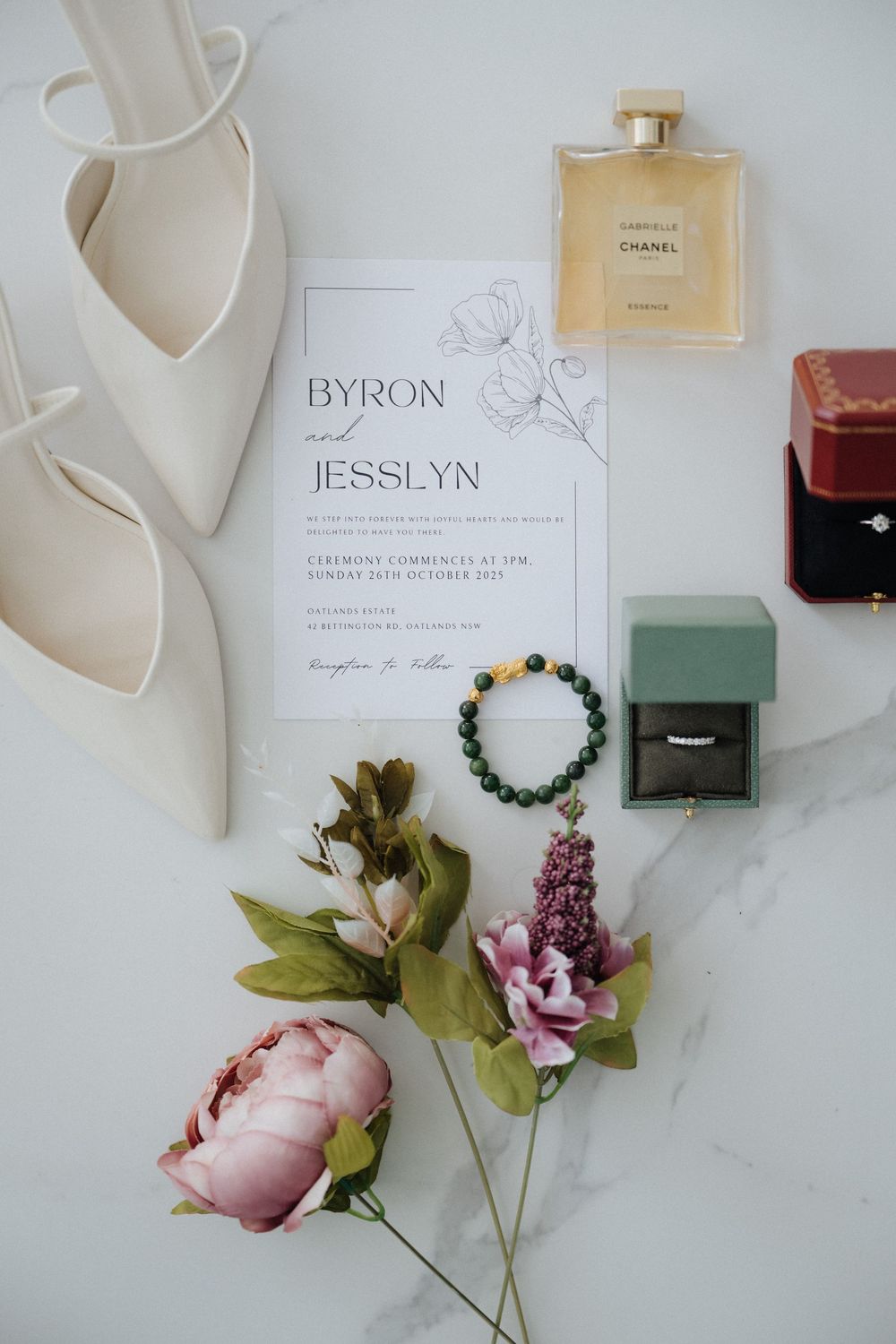 Elegant wedding flat lay featuring Byron and Jesslyn invitation, white heels, Chanel perfume, ring boxes, bracelet, and pink flowers.