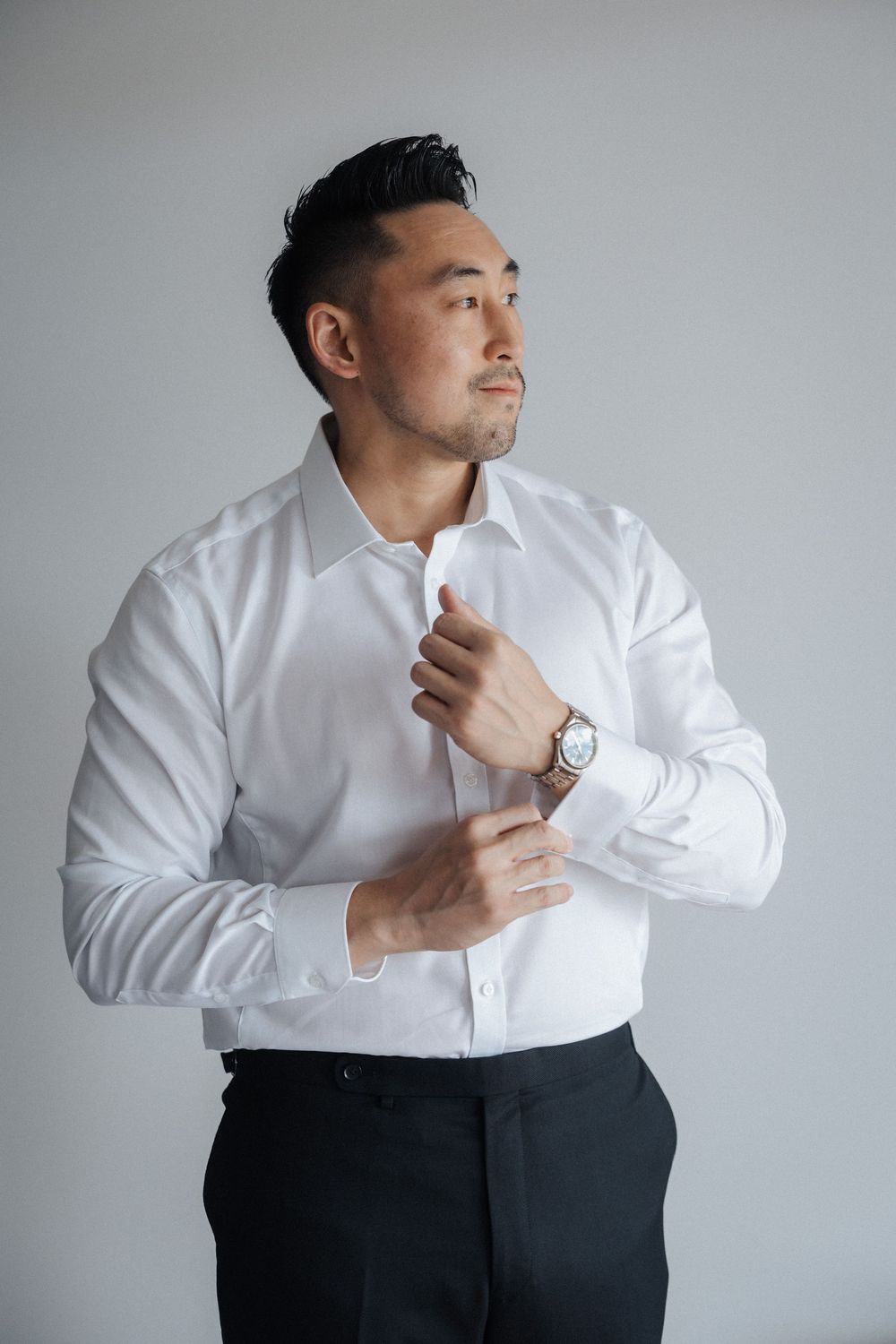 A well-dressed Asian man in a white dress shirt and black pants adjusts his collar while wearing a silver watch.