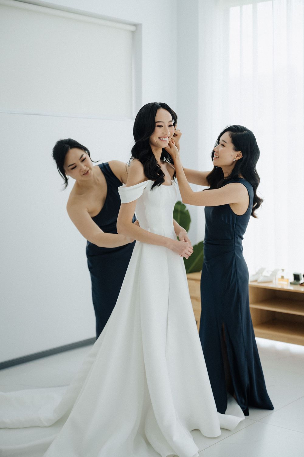 Bride in white off-shoulder gown being helped by two bridesmaids in navy dresses during wedding preparations.