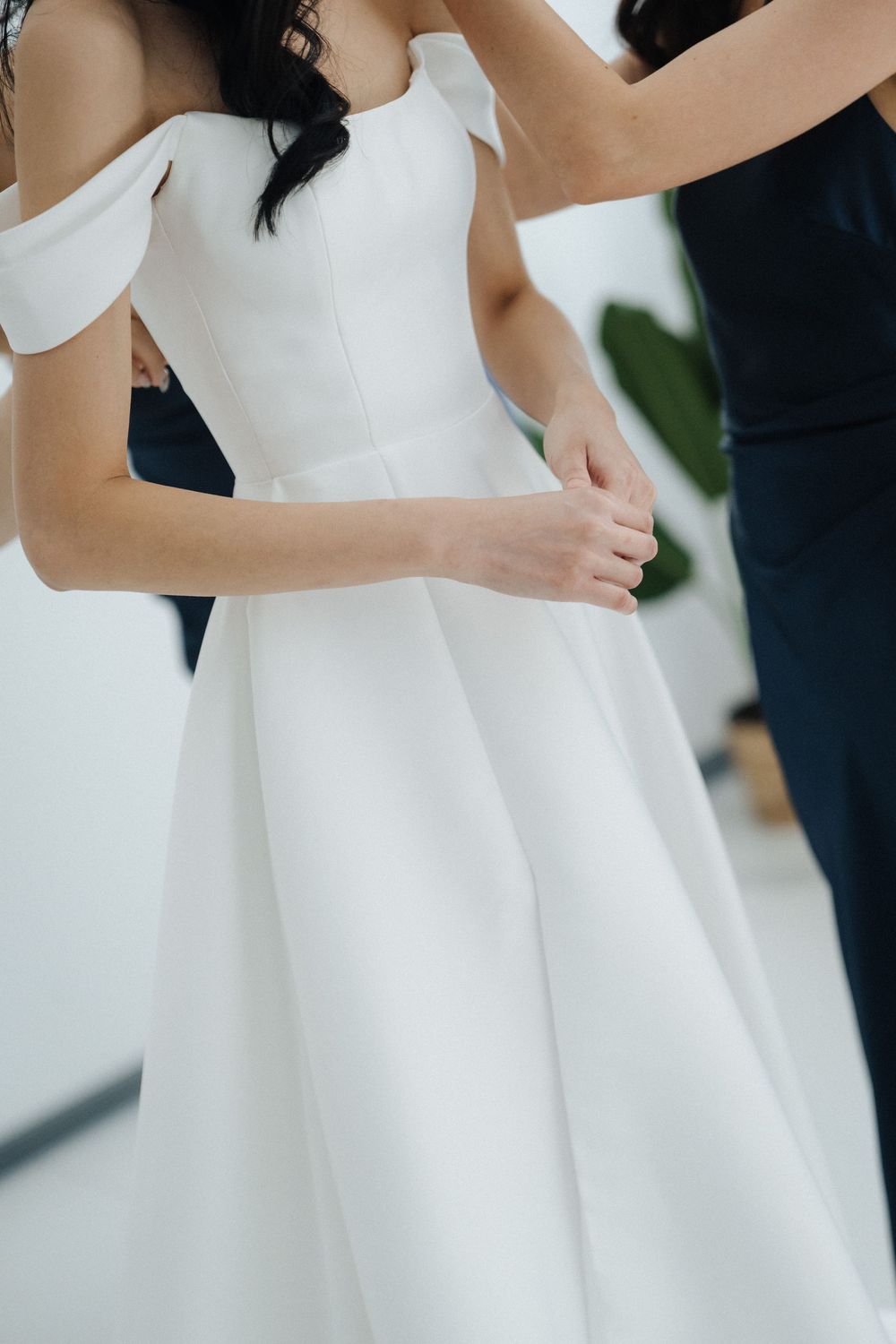 Bride in elegant off-shoulder white A-line wedding dress being assisted by bridesmaid in navy blue gown.