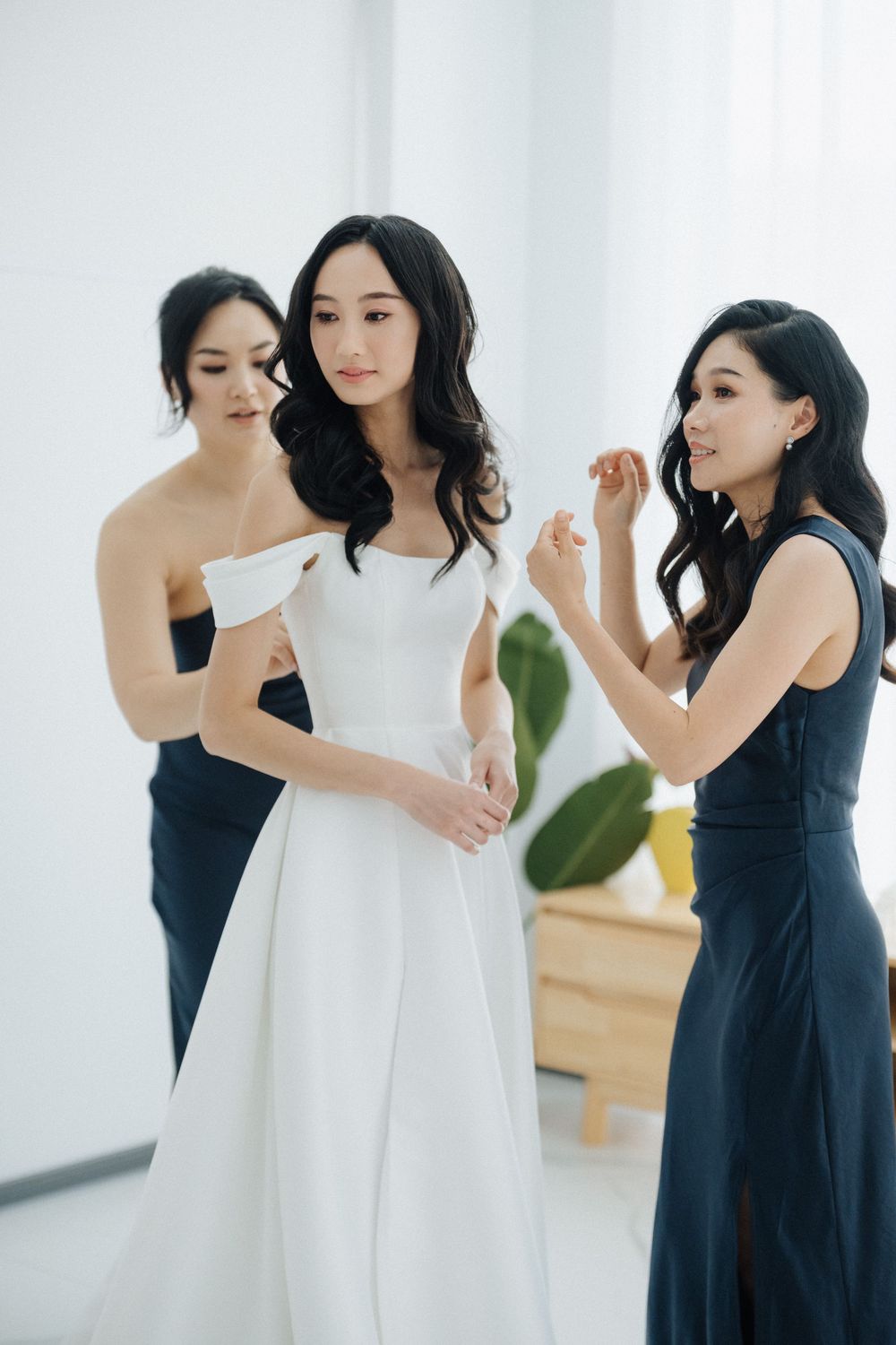 Bride in white off-shoulder gown assisted by two bridesmaids in navy dresses during wedding preparation.