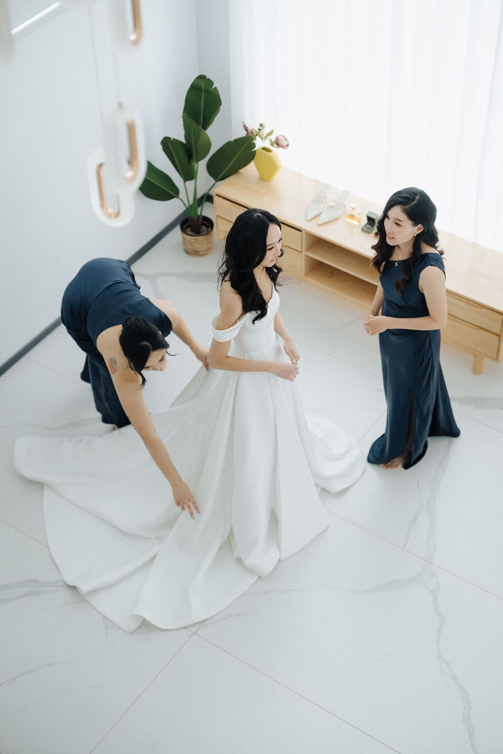 Bride in white gown being helped by two bridesmaids in navy dresses in a bright, modern room.