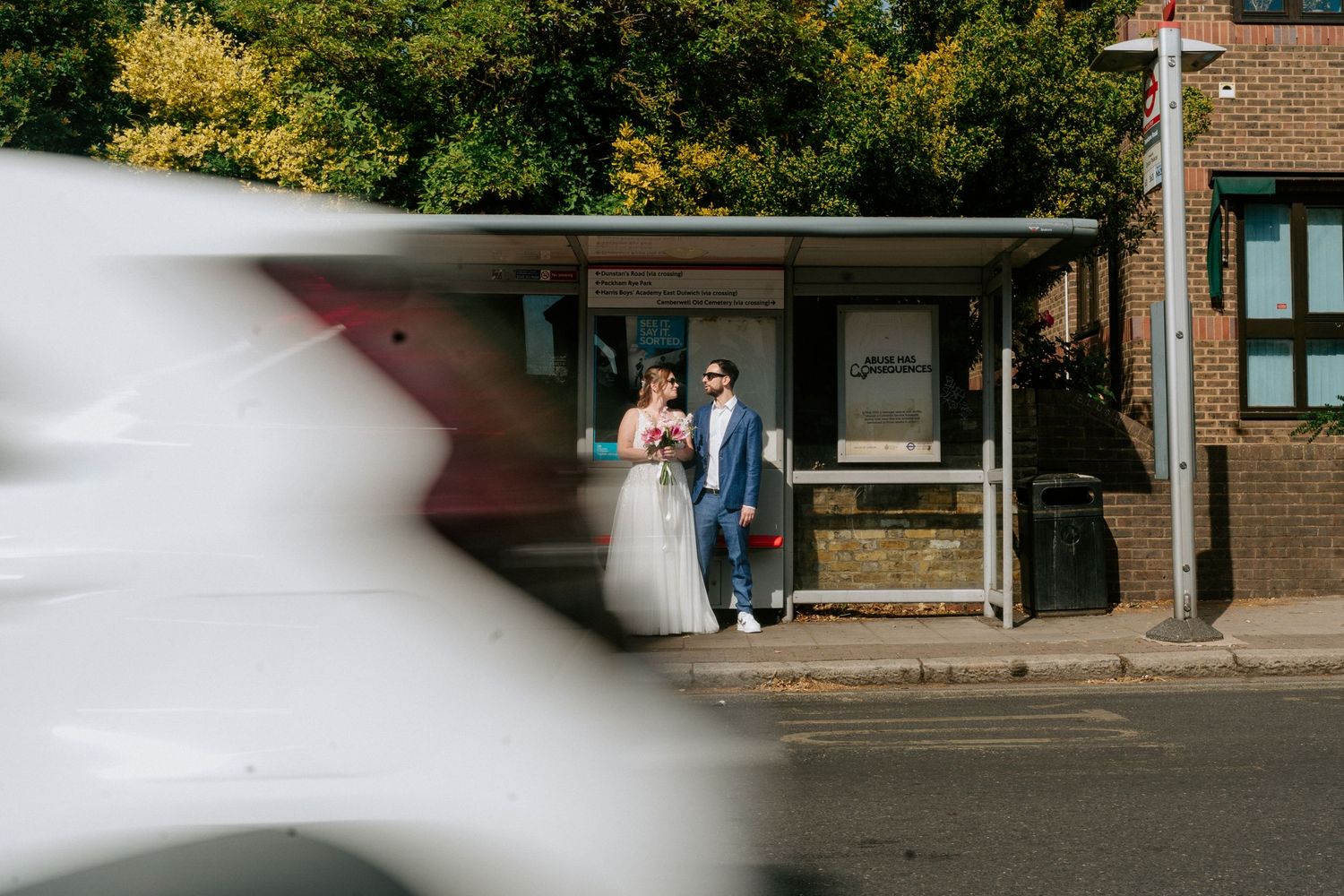 Bride and groom standing at a bus stop on a sunny street, with a white car passing in the foreground.