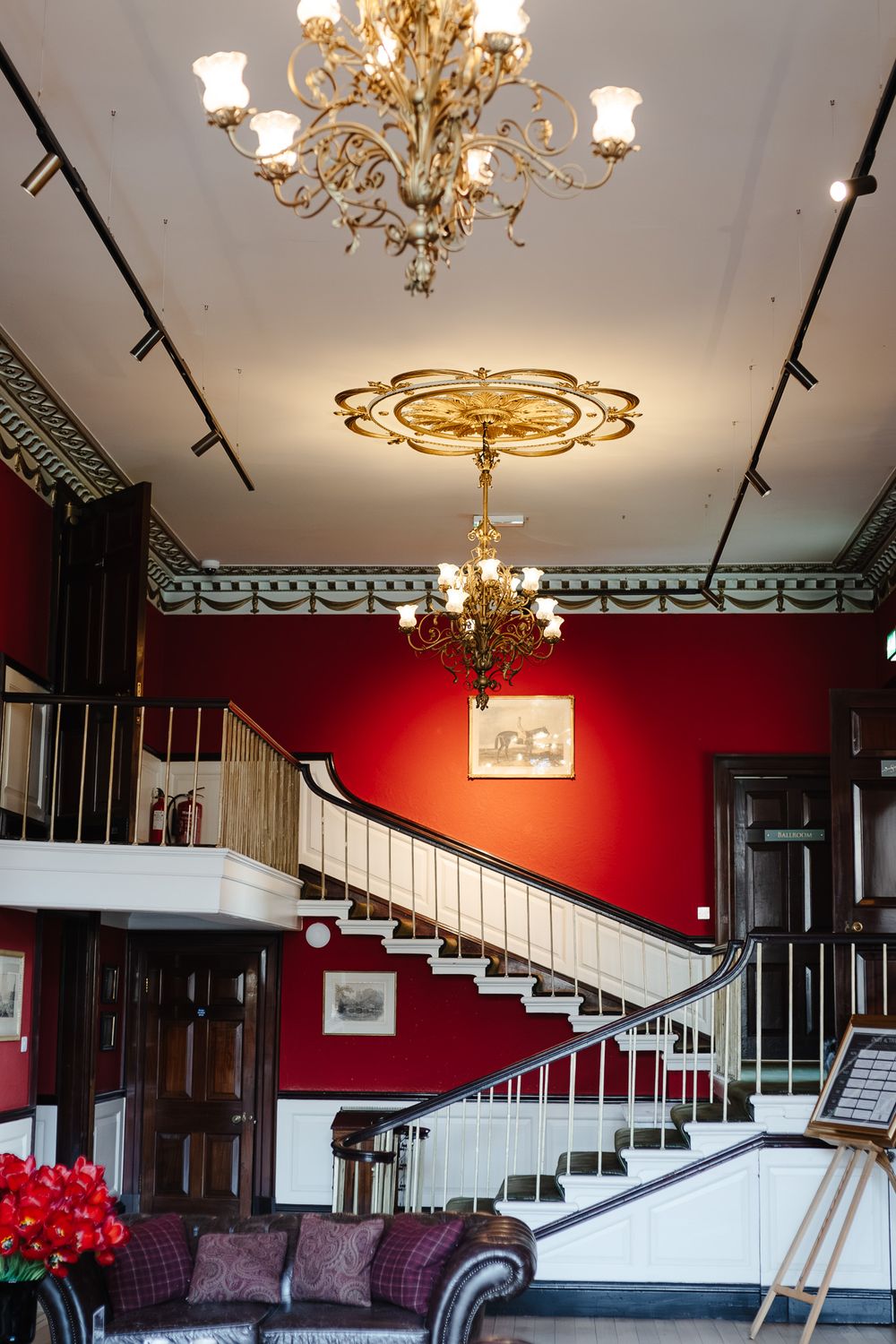 Grand hotel lobby with red walls, sweeping staircase, and ornate gold chandeliers hanging from ceiling.