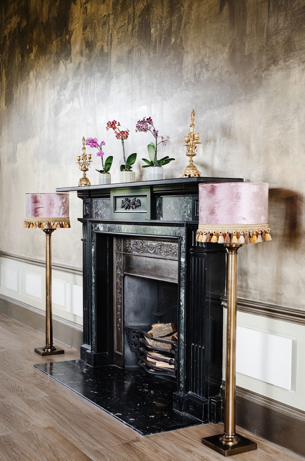 Elegant black marble fireplace with gold floor lamps, pink velvet shades, and orchids on mantel against textured gray wall.
