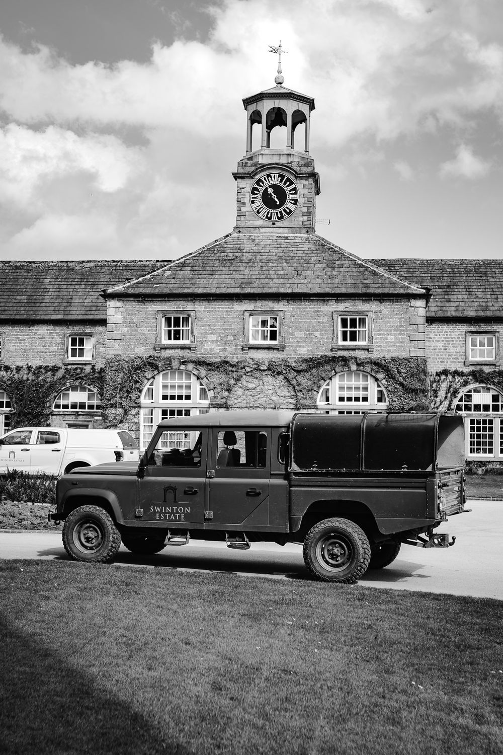 Black and white photo of a Land Rover Defender outside a historic stone estate building with clock tower.