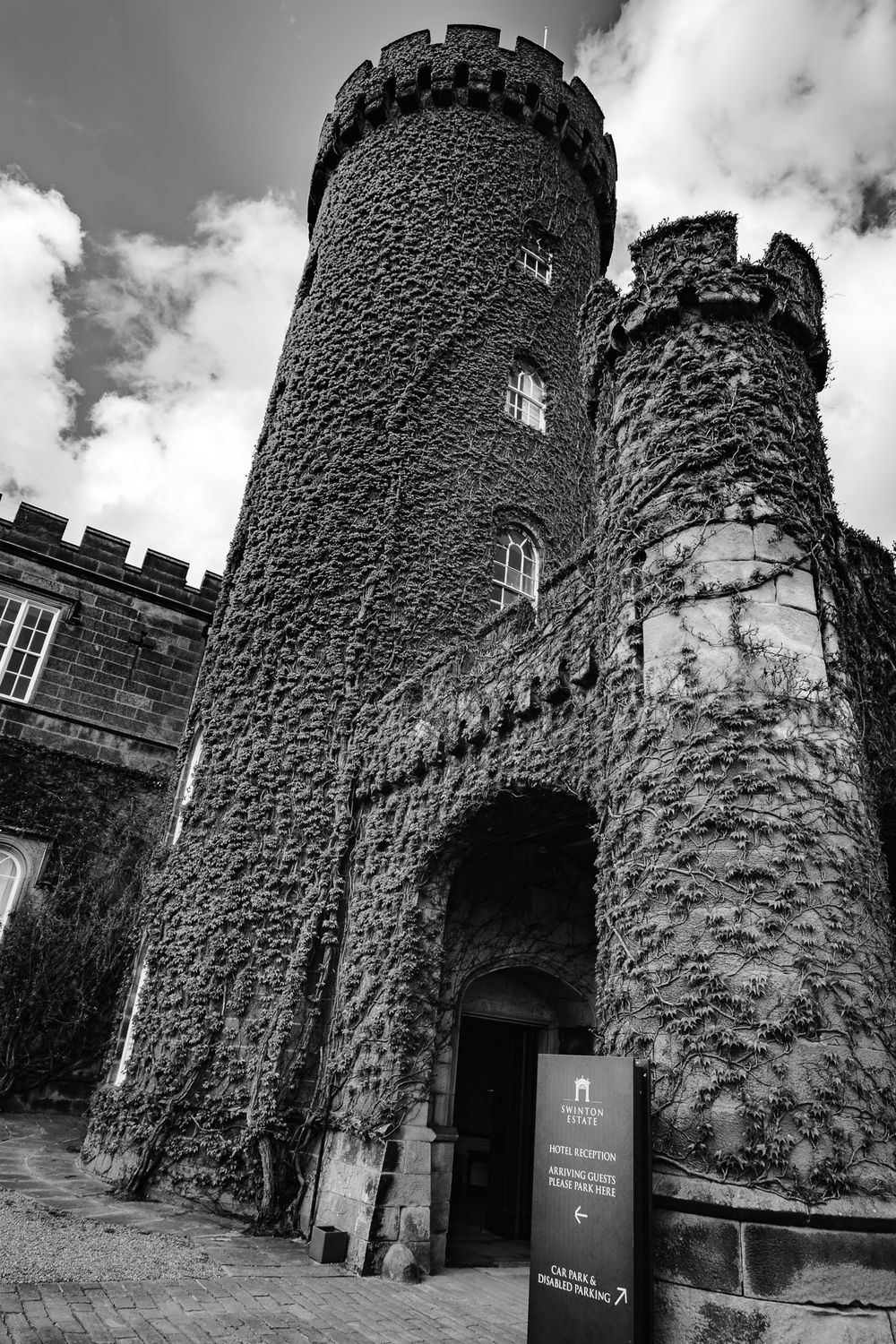 Black and white image of an ivy-covered castle tower entrance with a hotel reception sign on a cobblestone path.