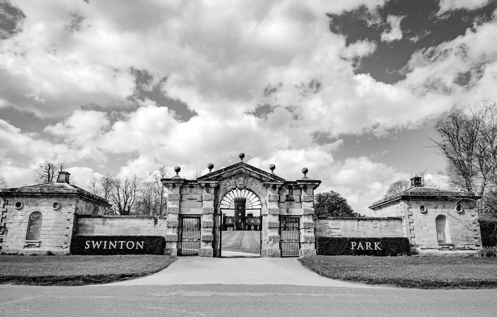 Black and white photo of Swinton Park entrance gates with stone pillars and cloudy sky.