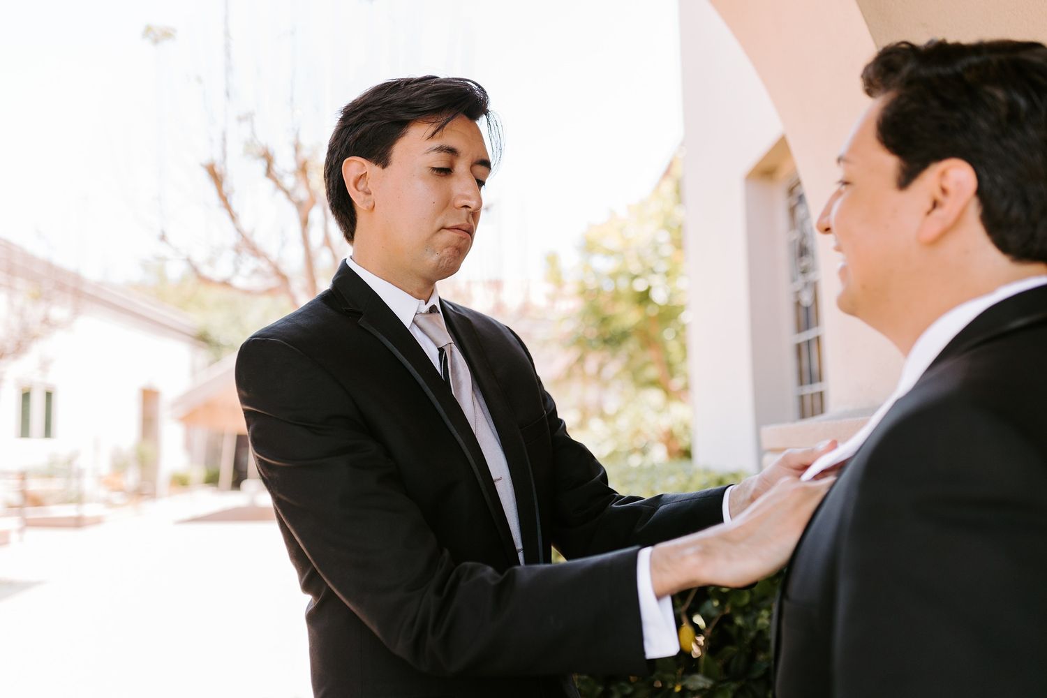 Two men in black suits adjusting ties outdoors at a formal event or wedding ceremony.