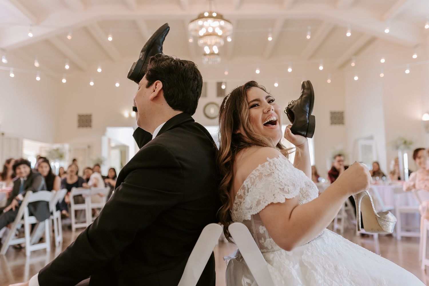 Bride and groom laughing during shoe game at elegant wedding reception in white venue with chandelier.