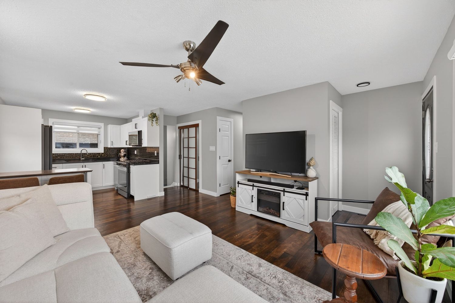 Open-concept living room with hardwood floors, ceiling fan, white sofa, and modern kitchen visible in background.