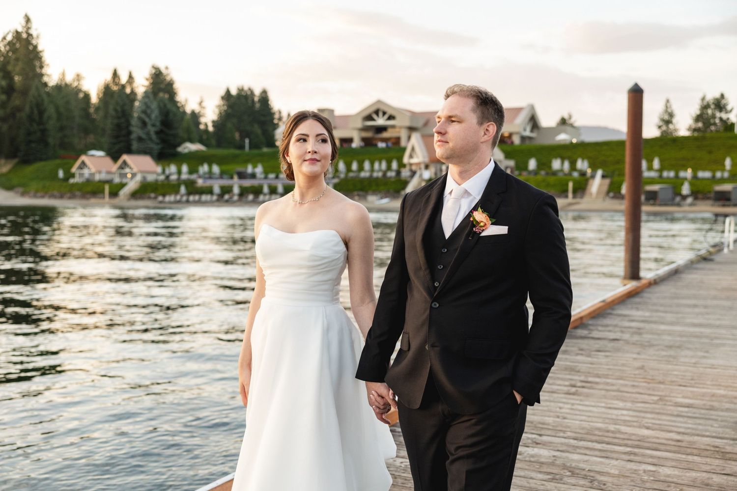 Bride and groom hold hands on a wooden dock by a lake at sunset, with lush green grounds in the background.
