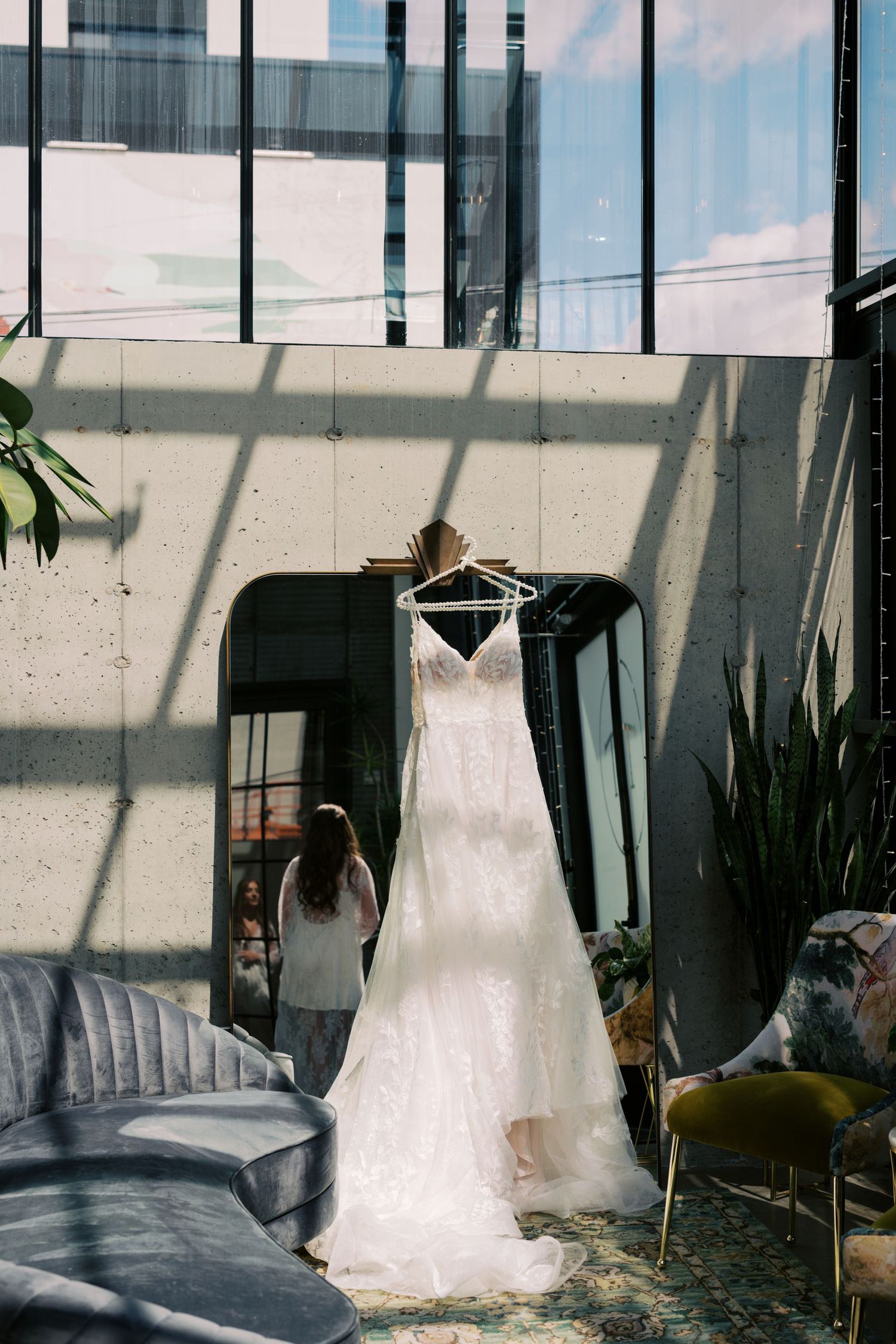 A white lace wedding dress hanging on a hanger in a modern venue with large windows and a woman standing nearby.