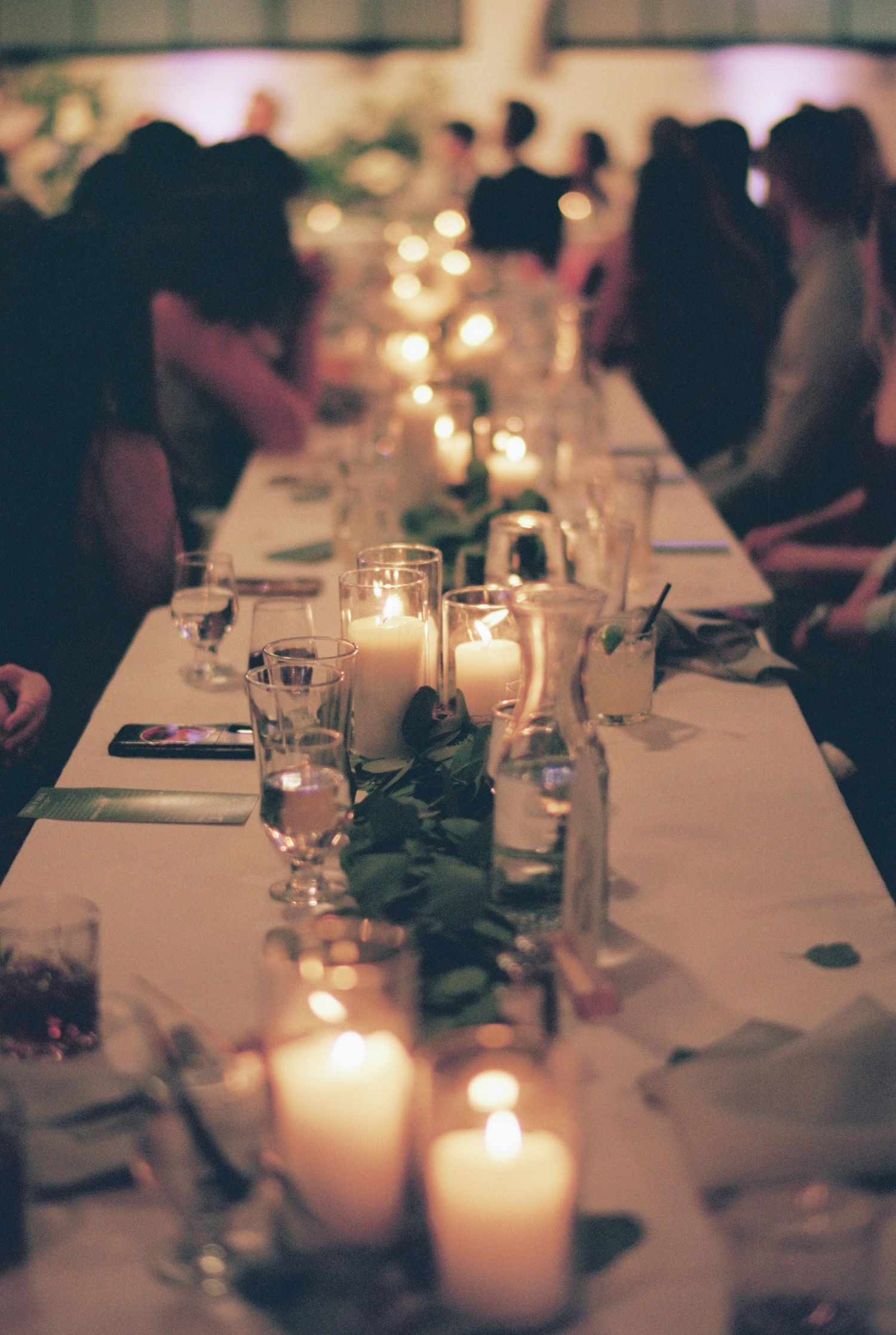 Elegant wedding reception long table adorned with glowing white pillar candles, greenery garland, and crystal glassware.