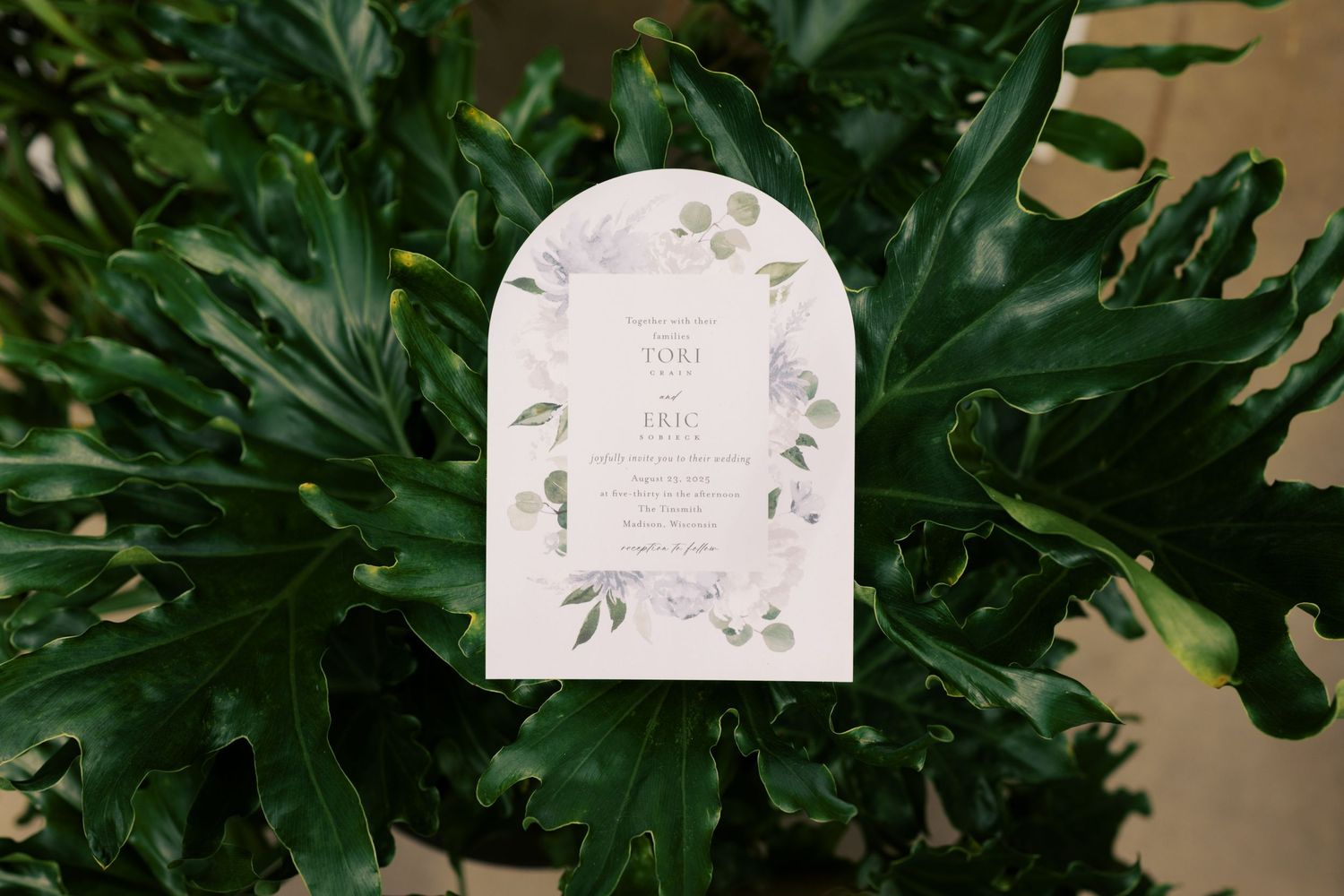Elegant arched wedding invitation with floral design displayed against lush tropical green leaves.