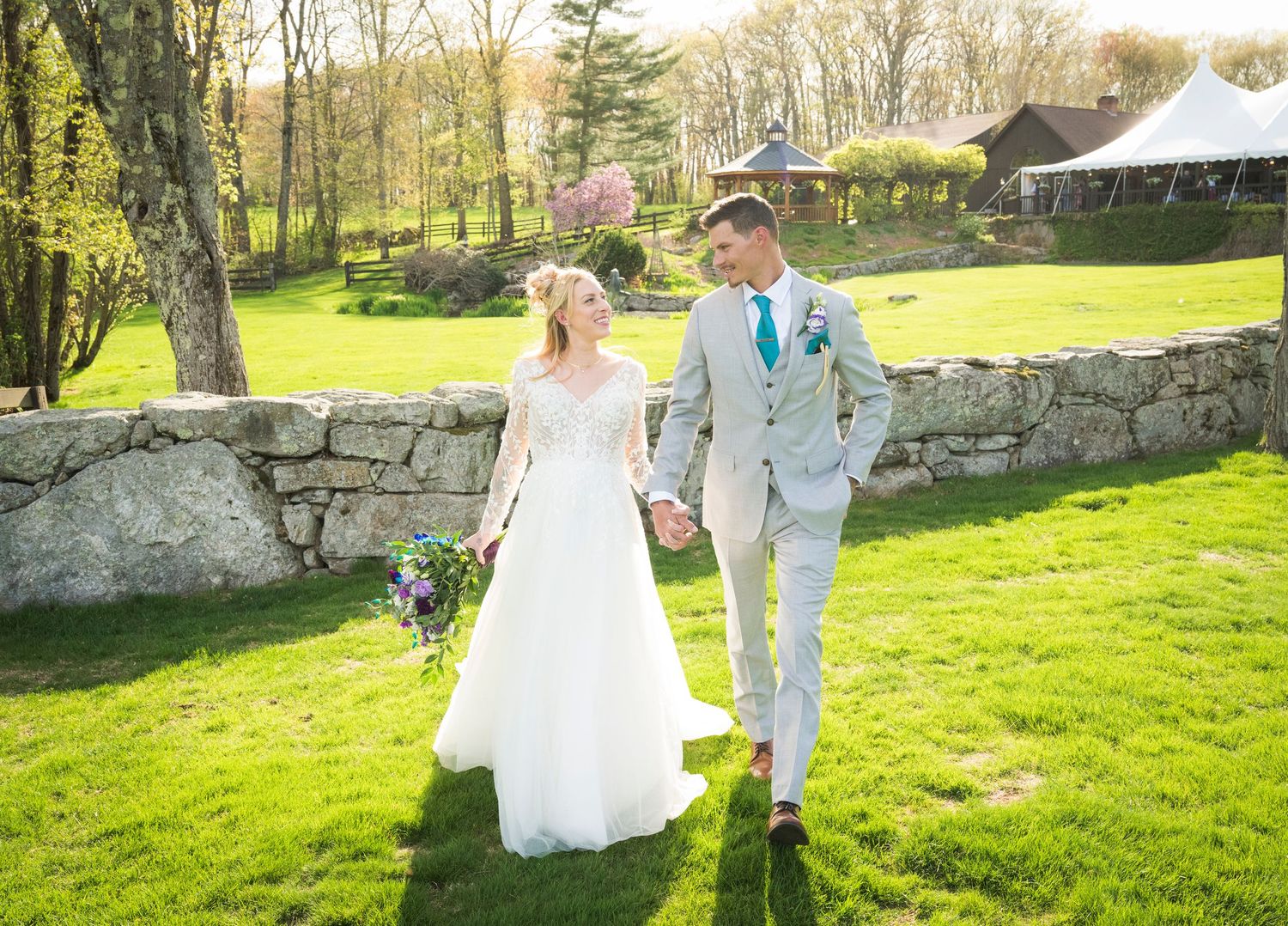 Bride and groom holding hands walking on green lawn beside stone wall at outdoor spring wedding venue.