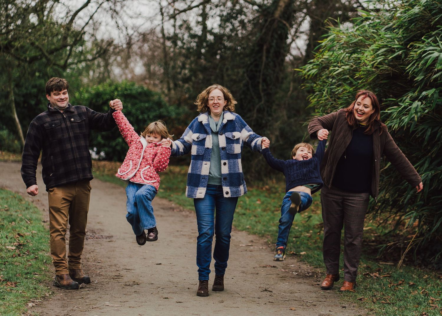 Family of five walking on a park path, laughing and swinging children by their hands outdoors.