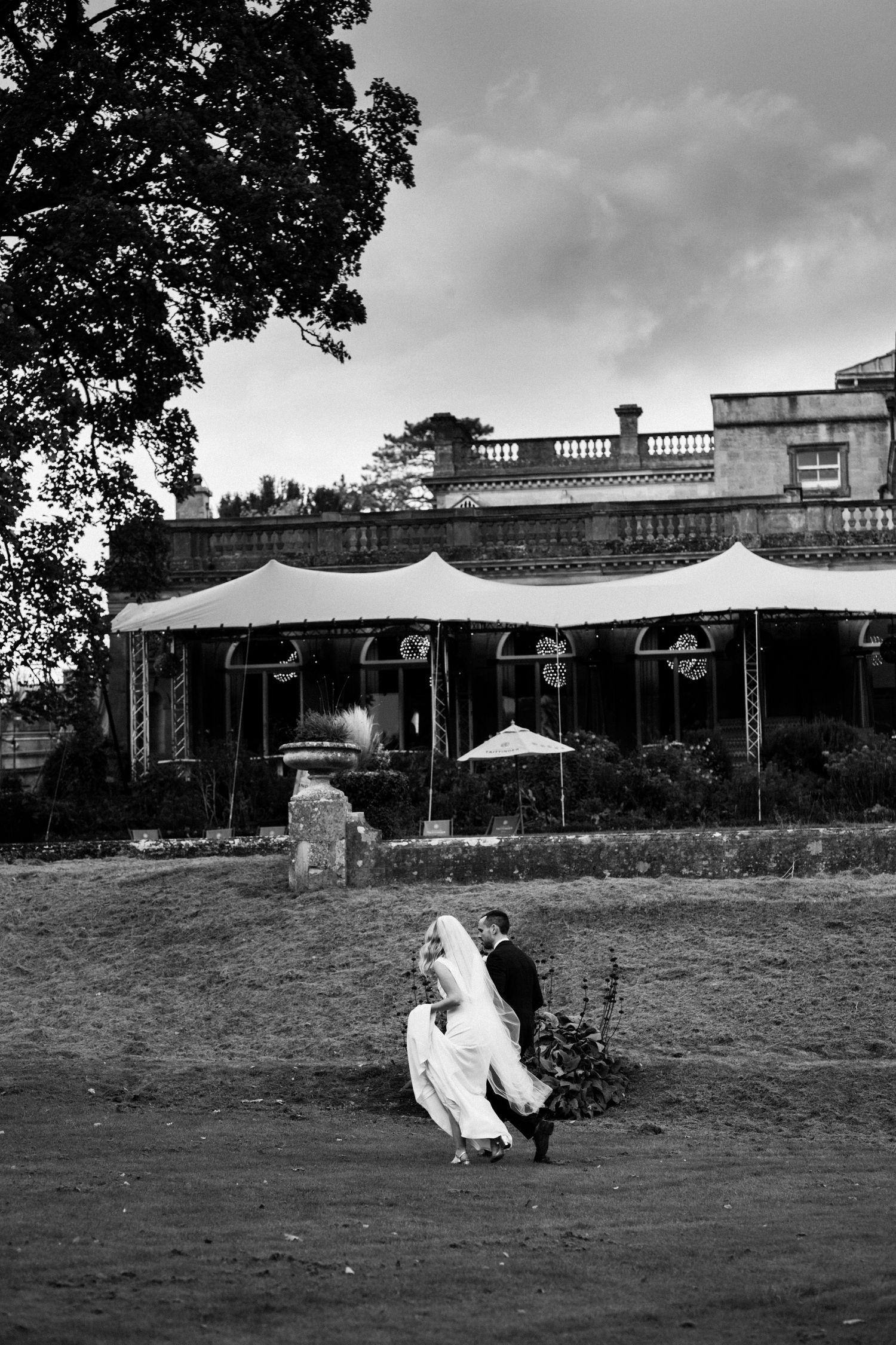 Black and white photo of a bride and groom walking outside a grand marquee venue on their wedding day.