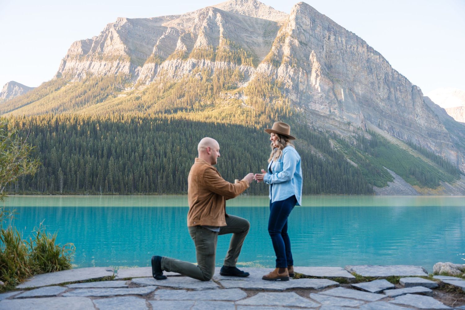 Man proposing to woman on one knee at turquoise Lake Louise with Rocky Mountain backdrop in Banff National Park.