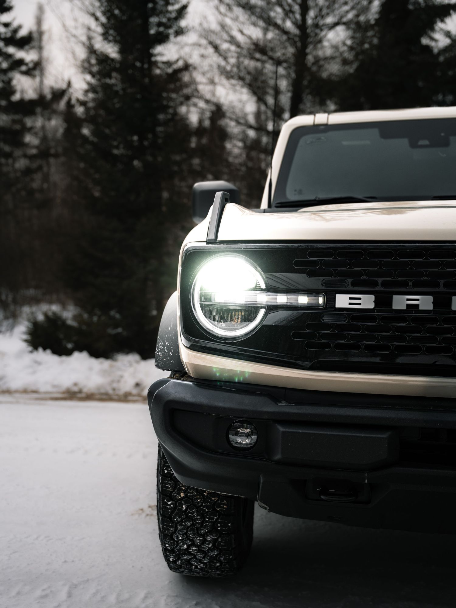 Close-up of a white Ford Bronco headlight glowing in a snowy winter setting with dark pine trees in background.