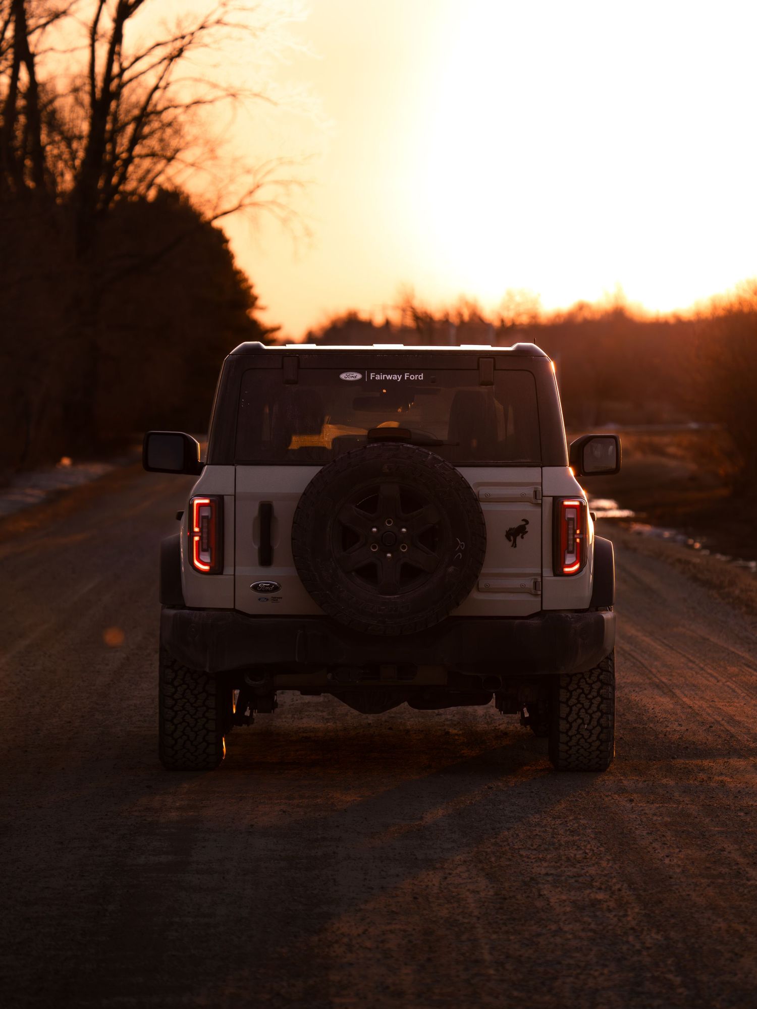 Rear view of a white Ford Bronco on a gravel road at sunset with glowing red taillights and a spare tire mount.
