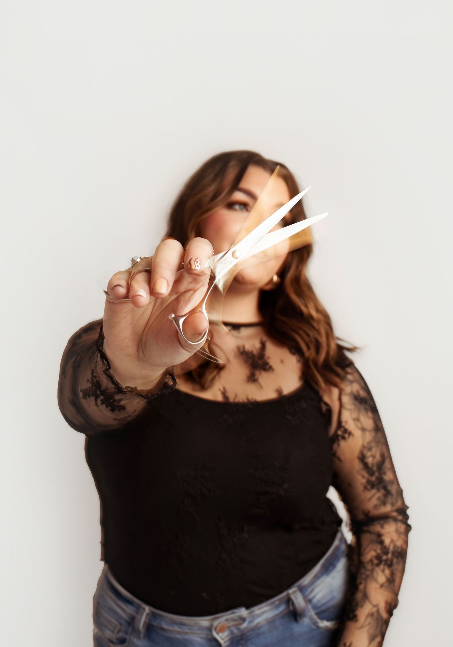 A woman in a black lace top holds scissors toward the camera in a professional hair stylist portrait.
