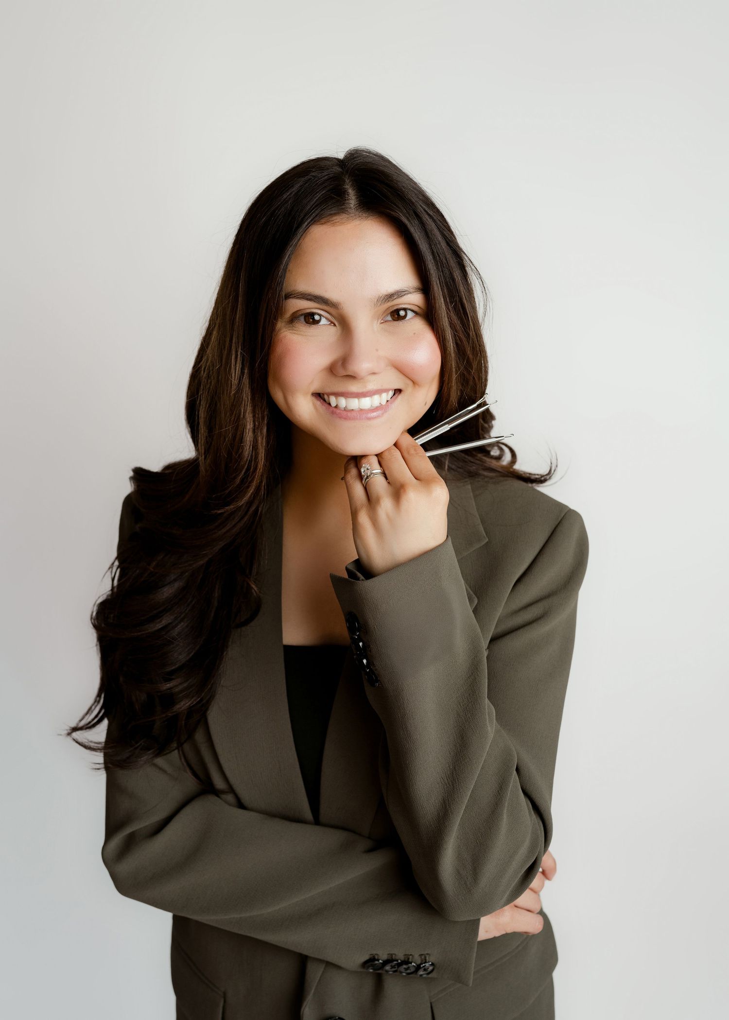 Professional woman in olive blazer smiling confidently while holding glasses against white background.