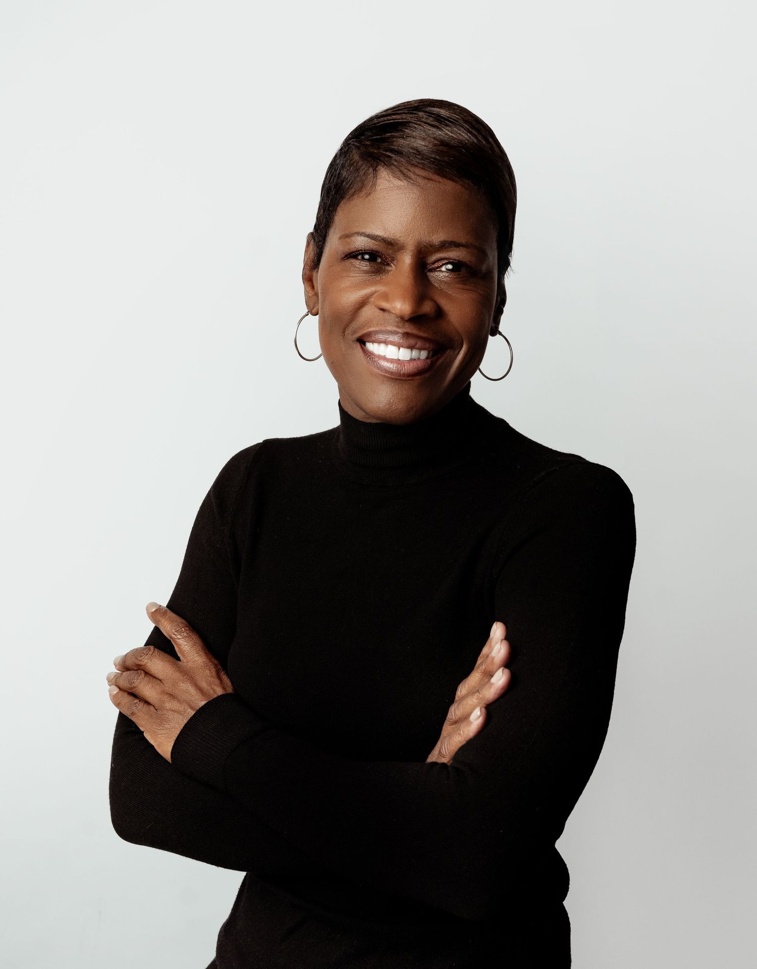 Confident woman in black turtleneck with arms crossed, smiling warmly against a light gray background.