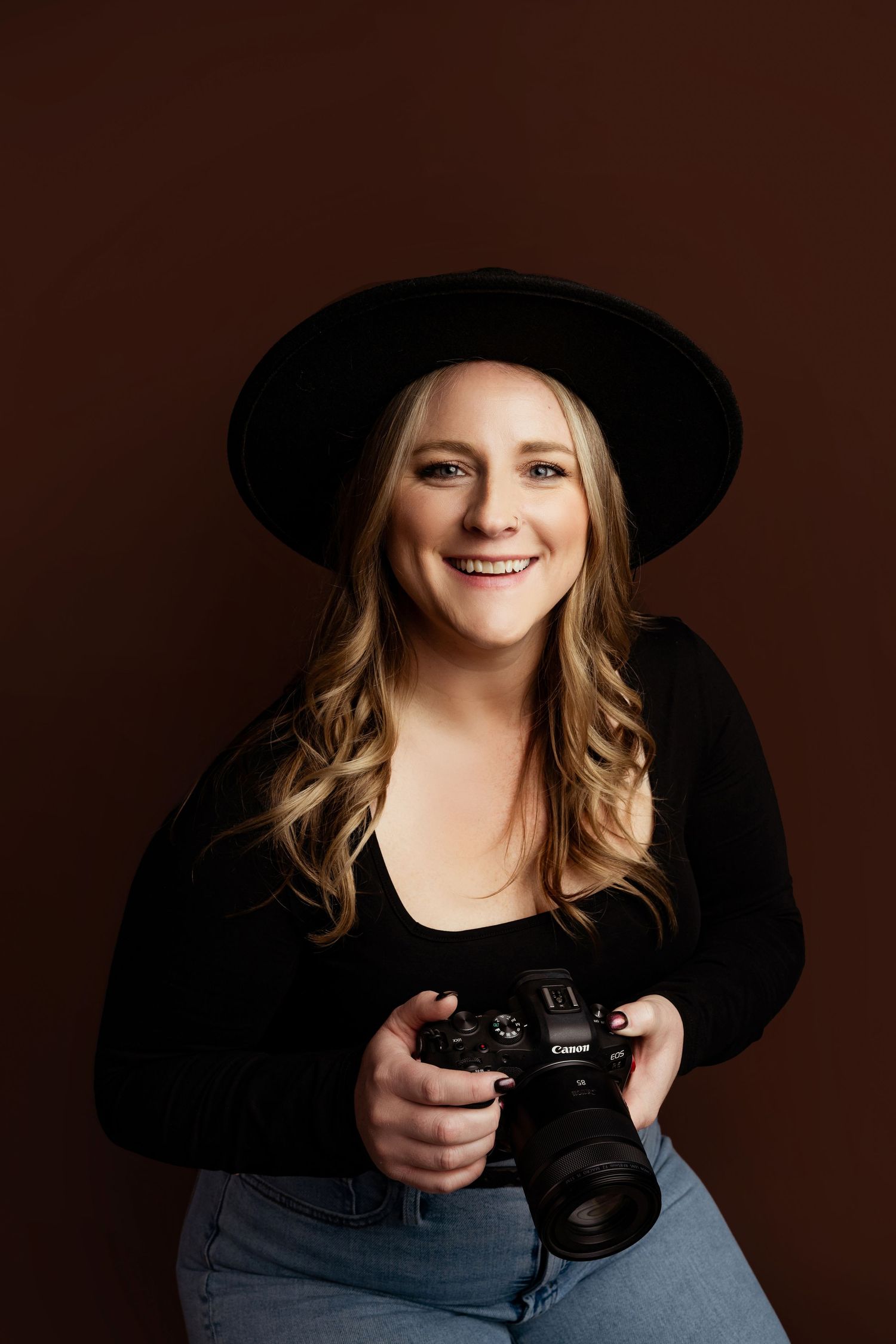 Smiling blonde woman in black hat and top holding a Canon camera against a brown studio background.