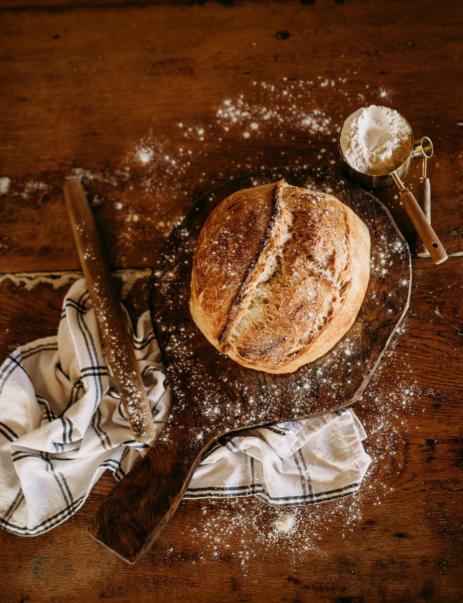 Rustic sourdough bread loaf dusted with flour on a wooden board with a bread knife and linen towel.